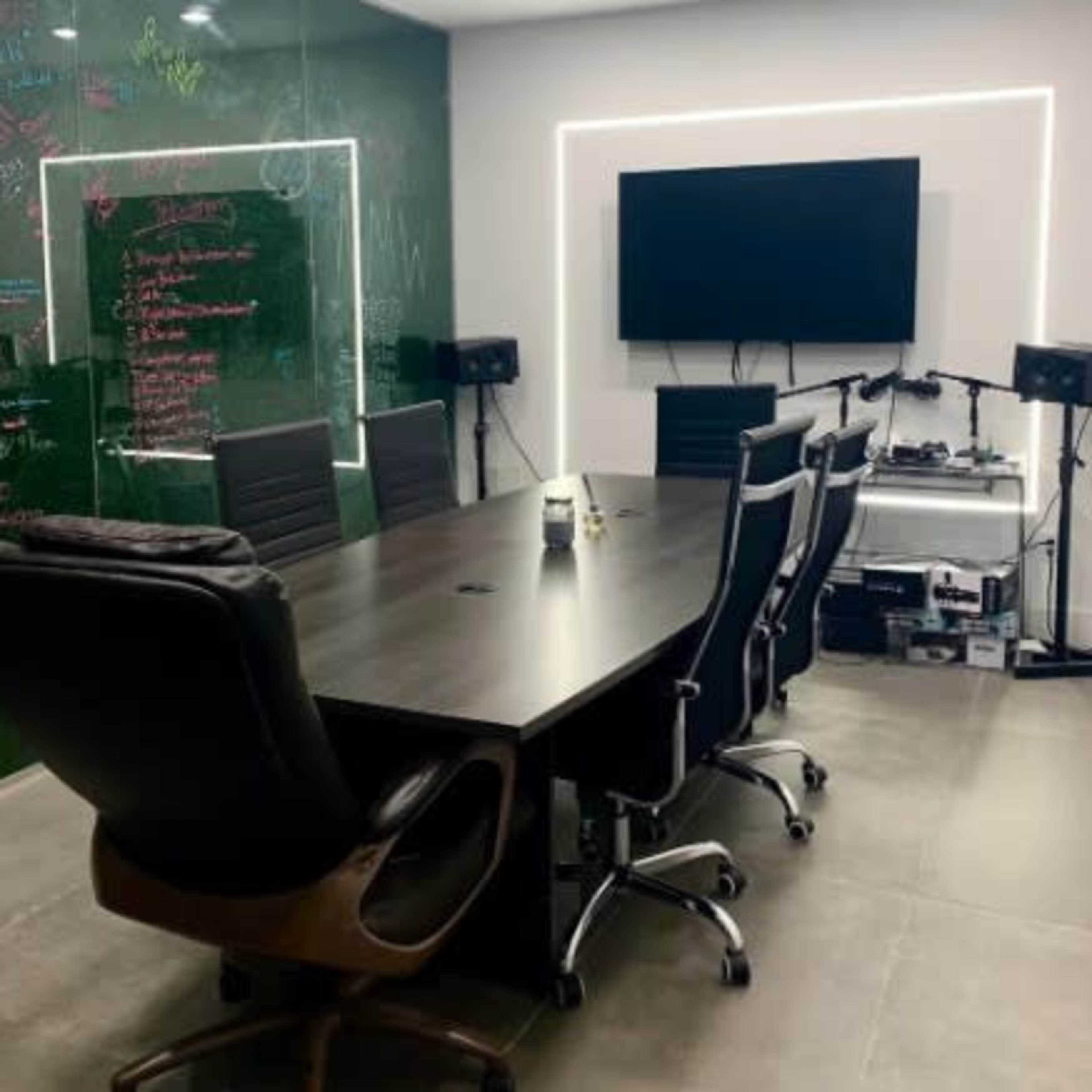 A modern conference room features a large table, ergonomic chairs, a television on the wall, and a green chalkboard with notes and diagrams.