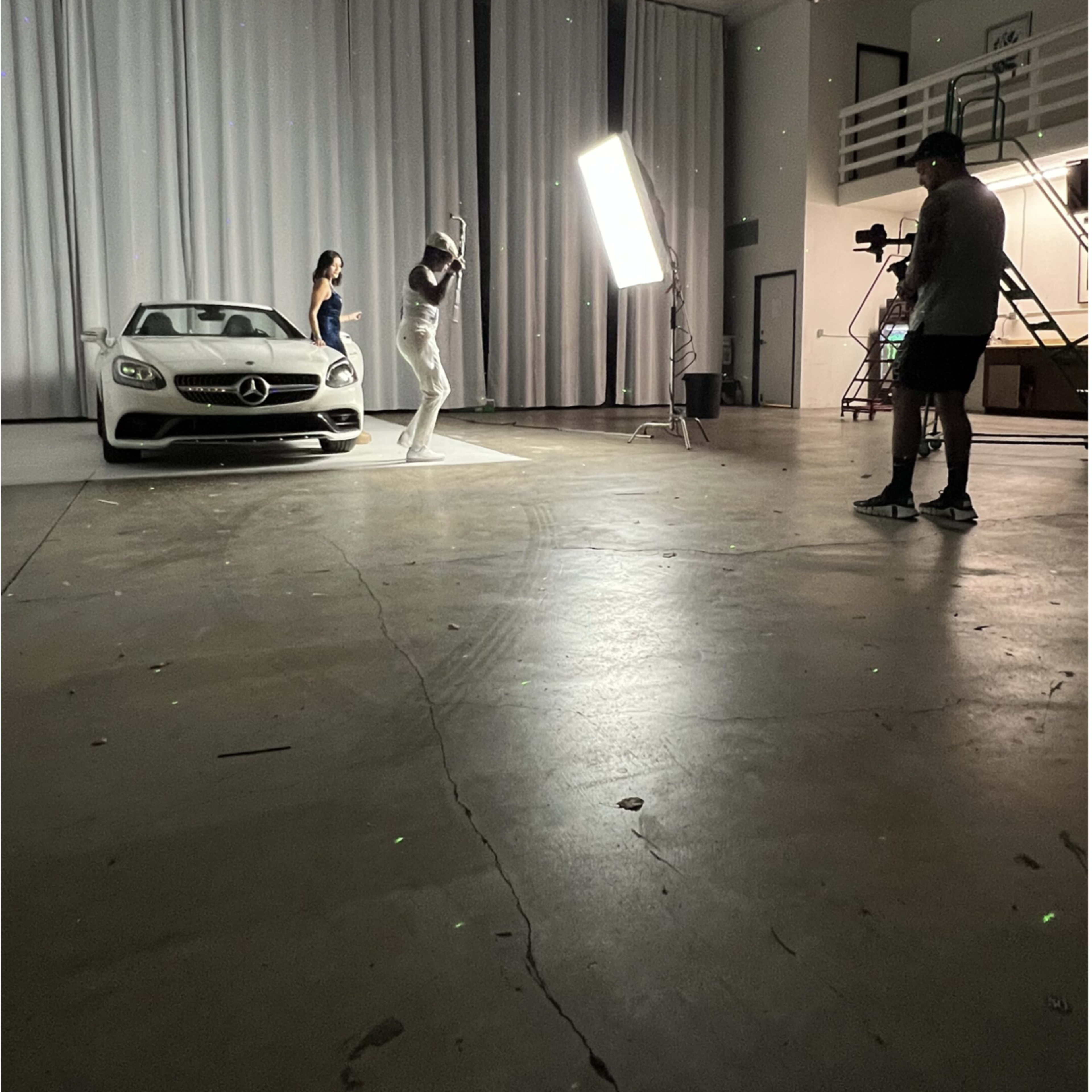 A man and a woman pose in front of a white Mercedes-Benz in a studio with draped curtains and a bright lighting setup.