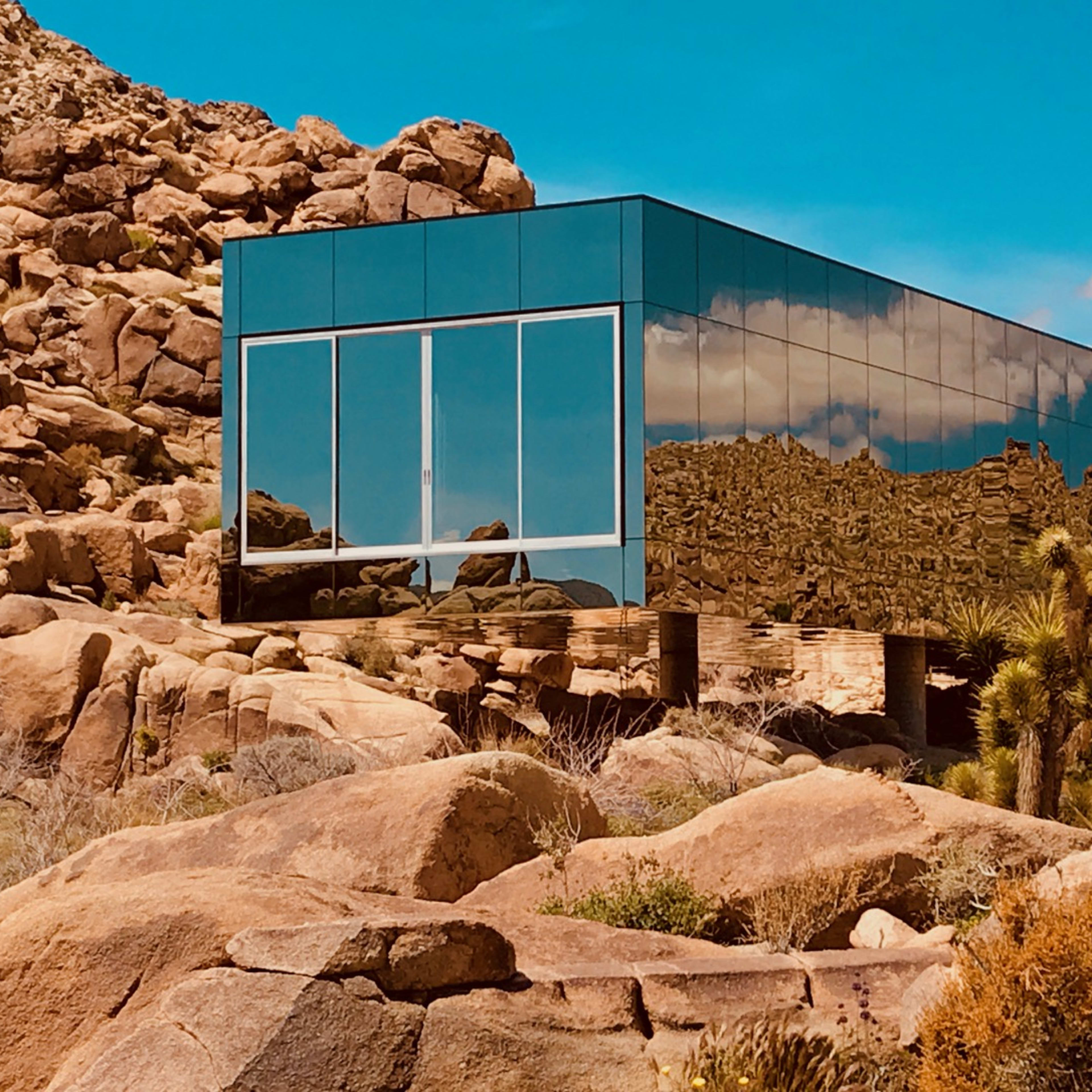 A modern glass building reflects the surrounding rocky landscape and desert vegetation under a blue sky.