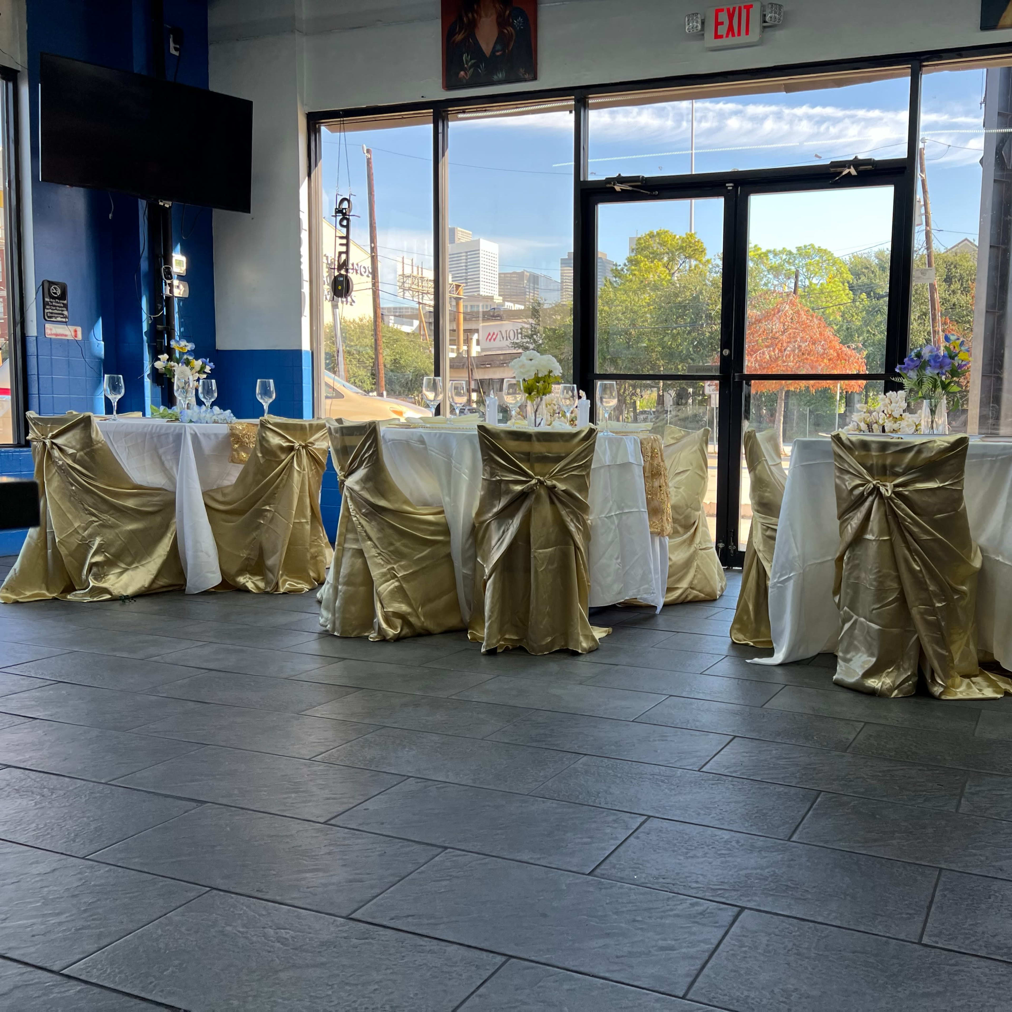 A set of elegantly decorated tables with gold and white tablecloths arranged near large windows in a brightly lit indoor space.