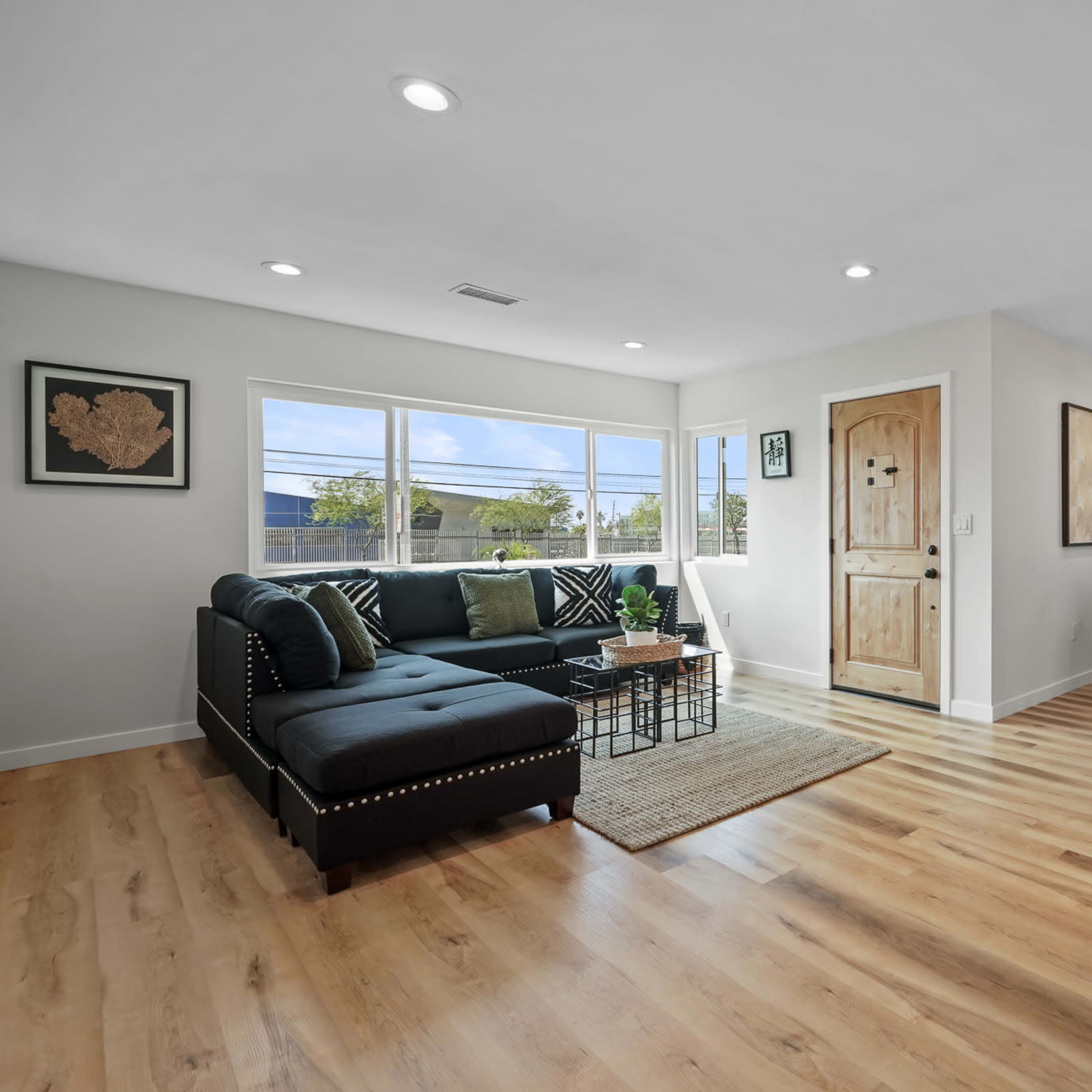 A modern living room with a black sectional sofa, large windows, a natural wood door, and a dining area featuring a small table and chairs.
