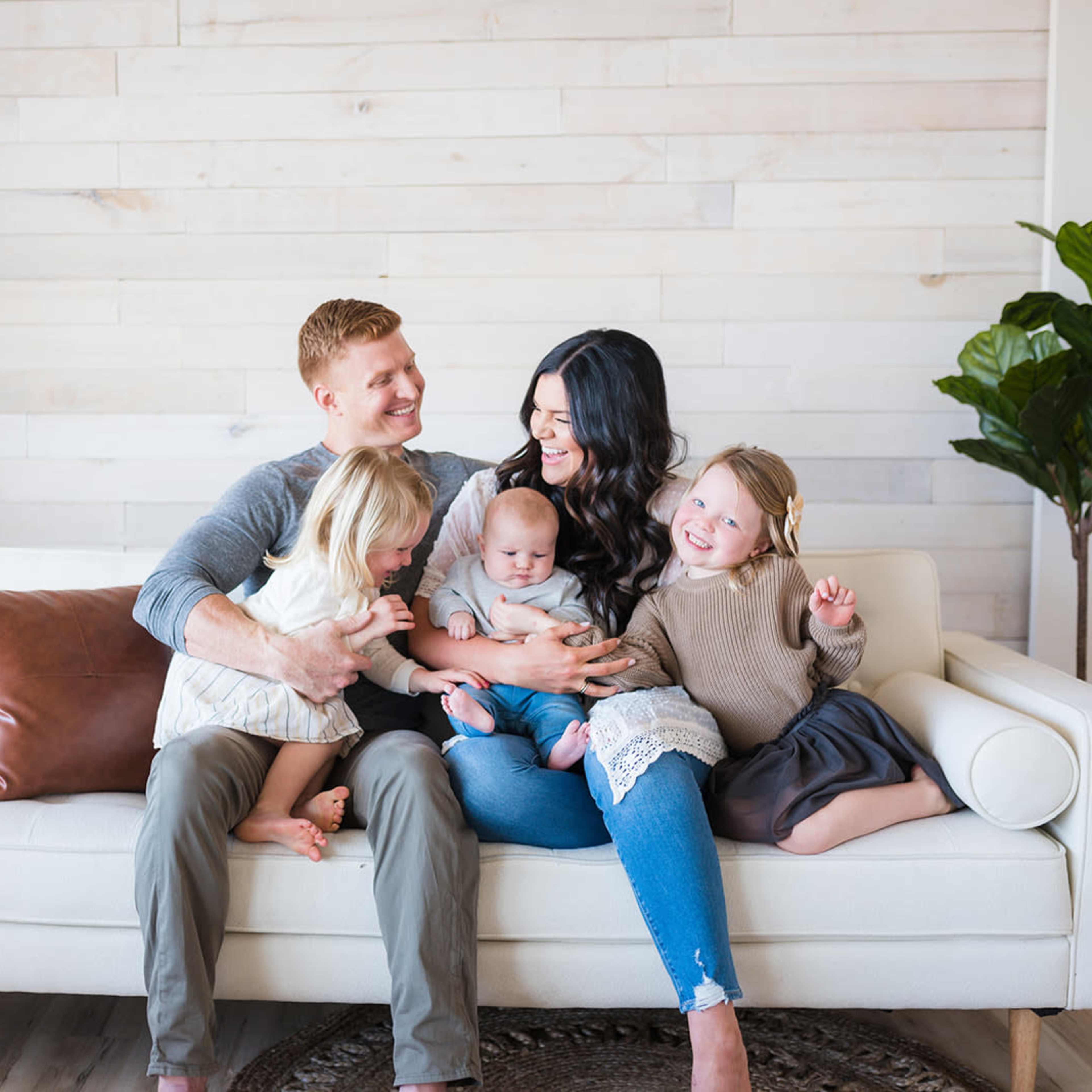 A family of five sits together on a white sofa in a bright room with wood paneling, smiling and interacting with each other.