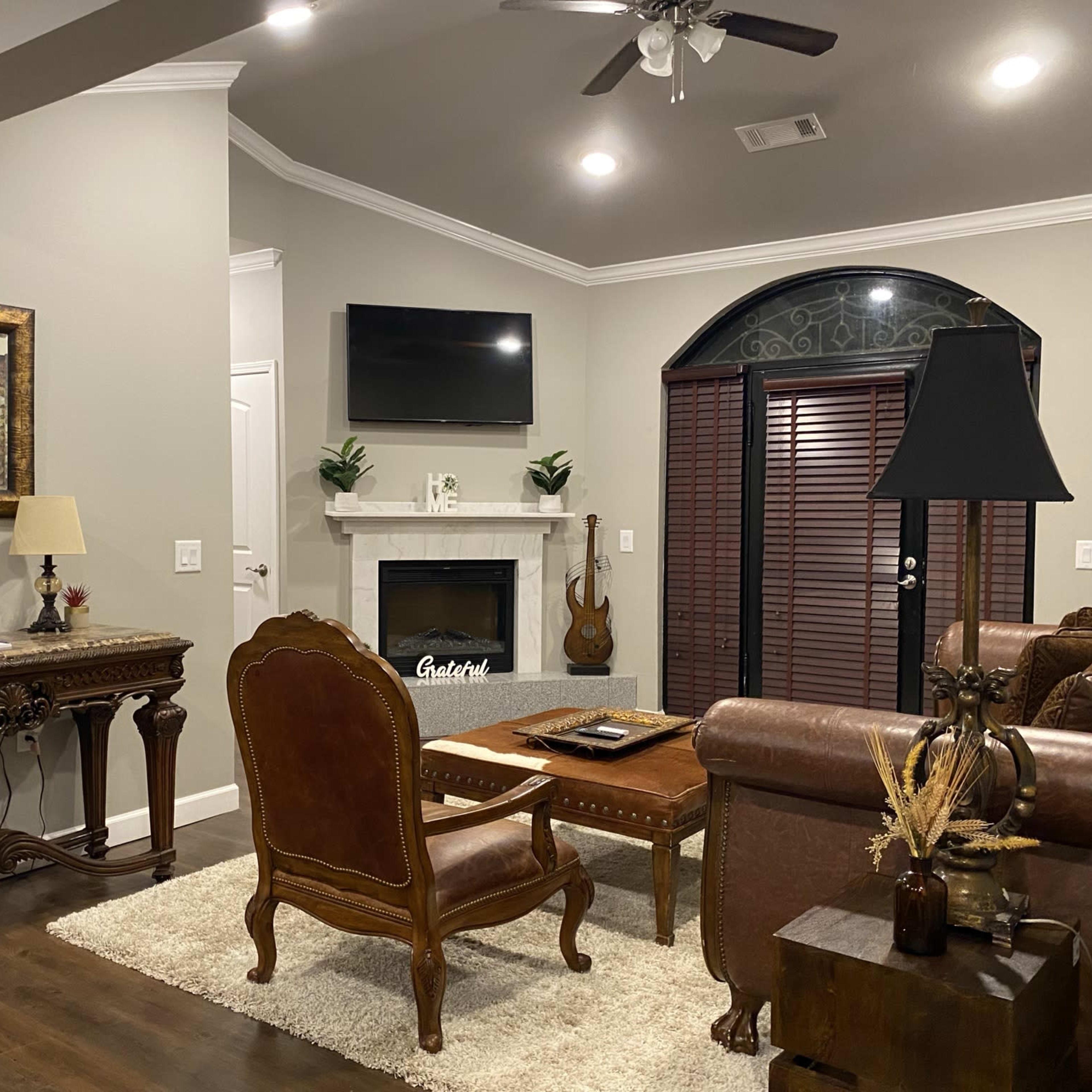 A spacious living room featuring a brown leather sofa, a wooden console table with decorative items, a fireplace with a television above it, and a chair positioned nearby.