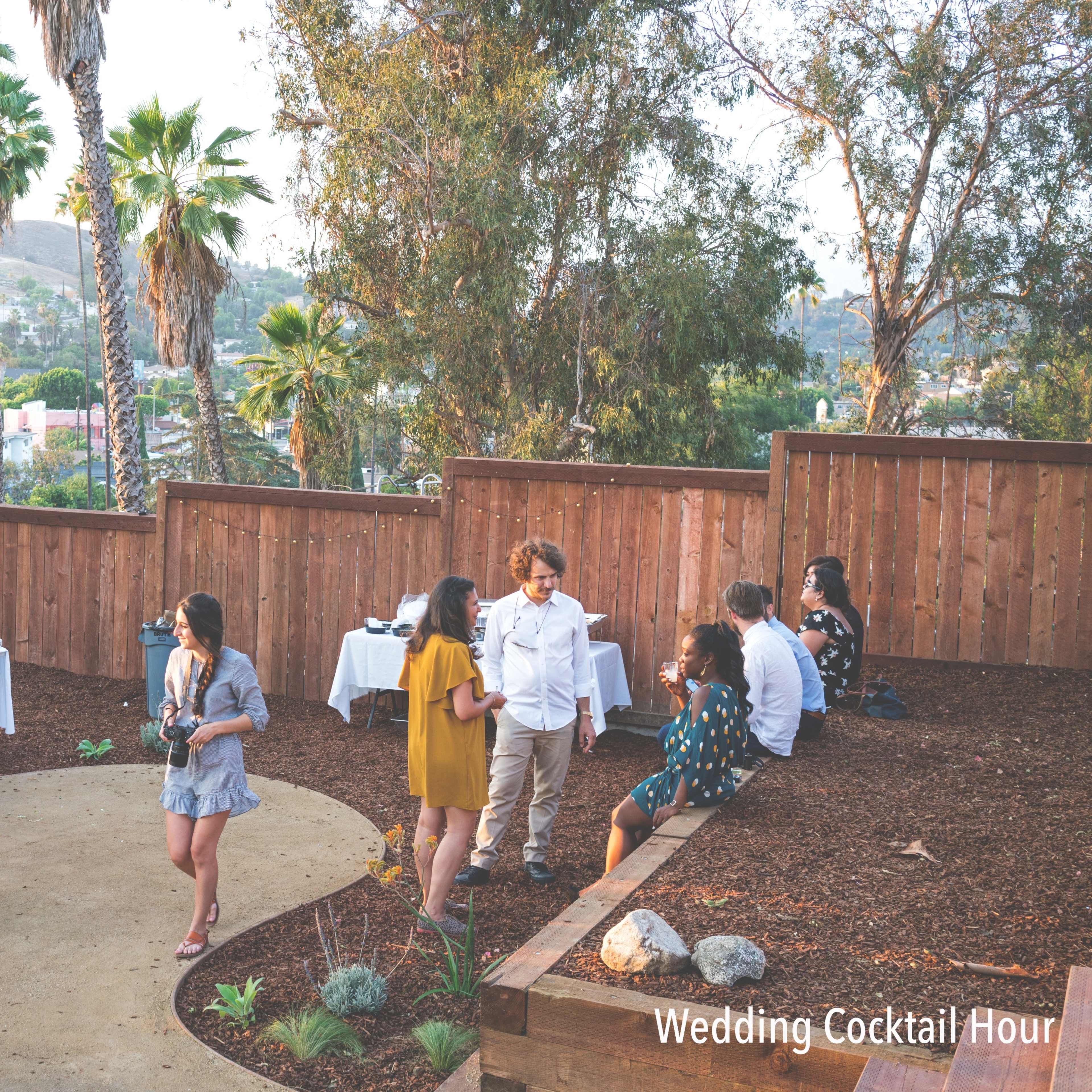 A group of guests enjoys a wedding cocktail hour in an outdoor setting surrounded by palm trees and wooden fences.