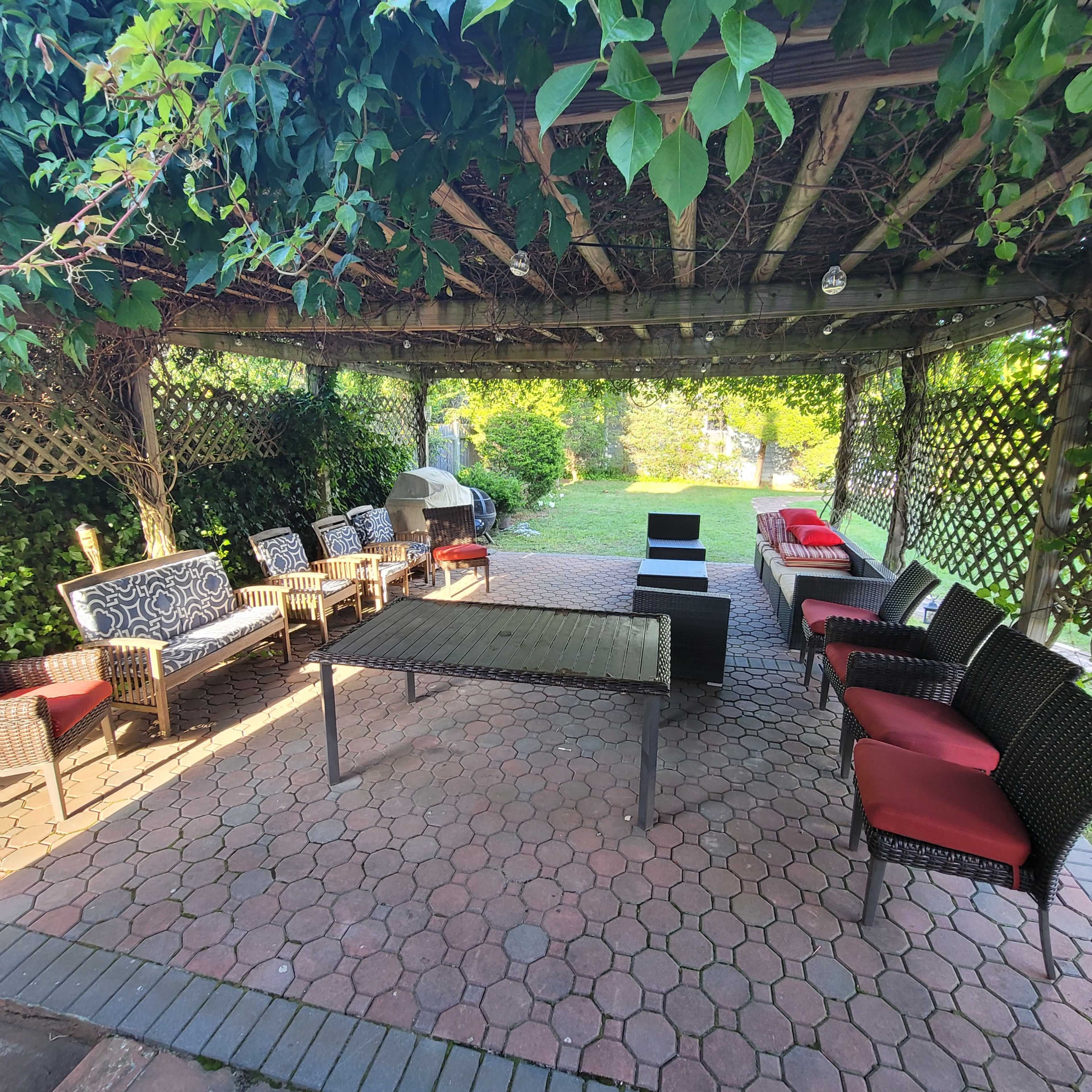 A covered patio area with several chairs and a table arranged around a stone-paved floor, surrounded by greenery.