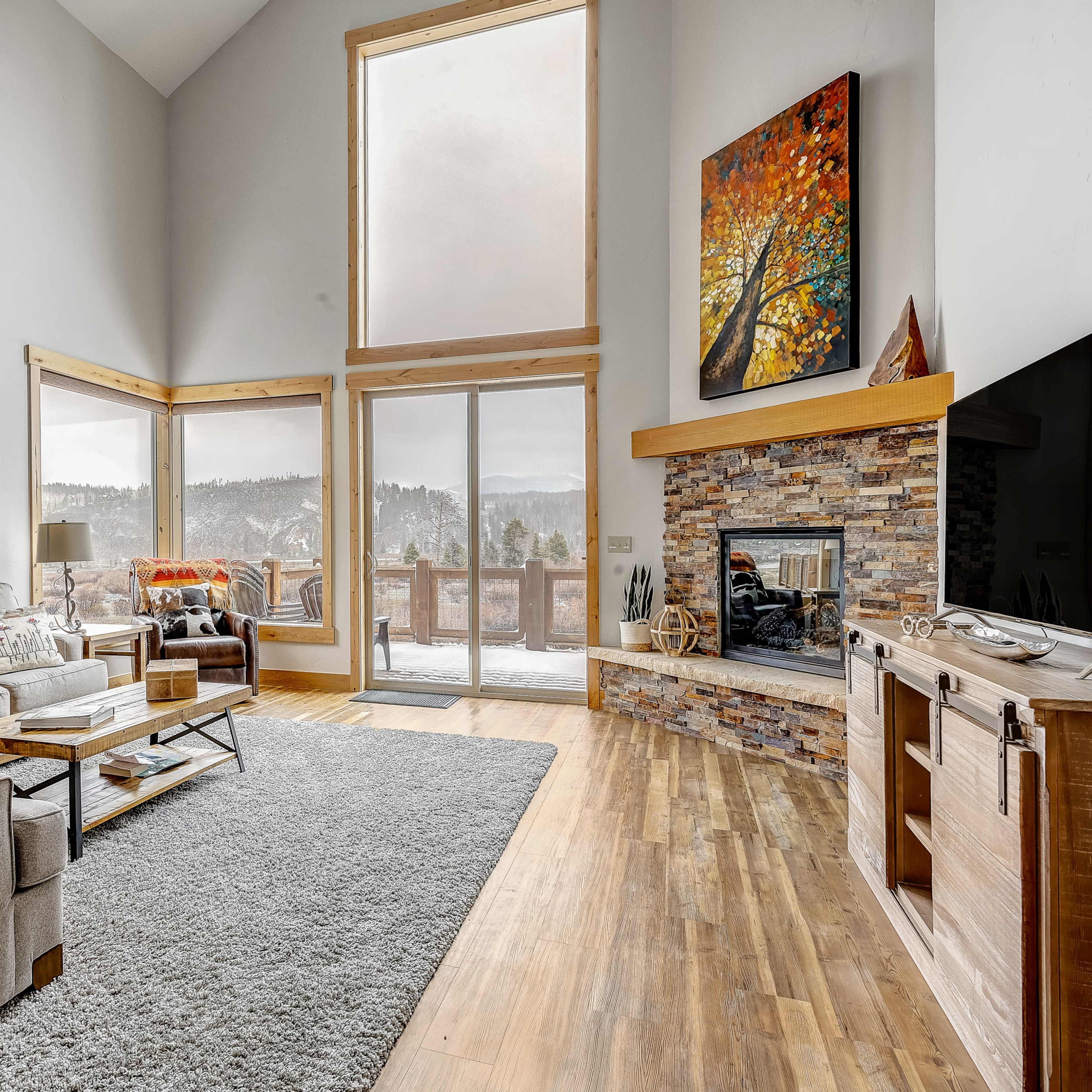 A spacious living room features a large window with a mountain view, a comfortable sectional sofa, a stone fireplace, and a television on a wooden cabinet.