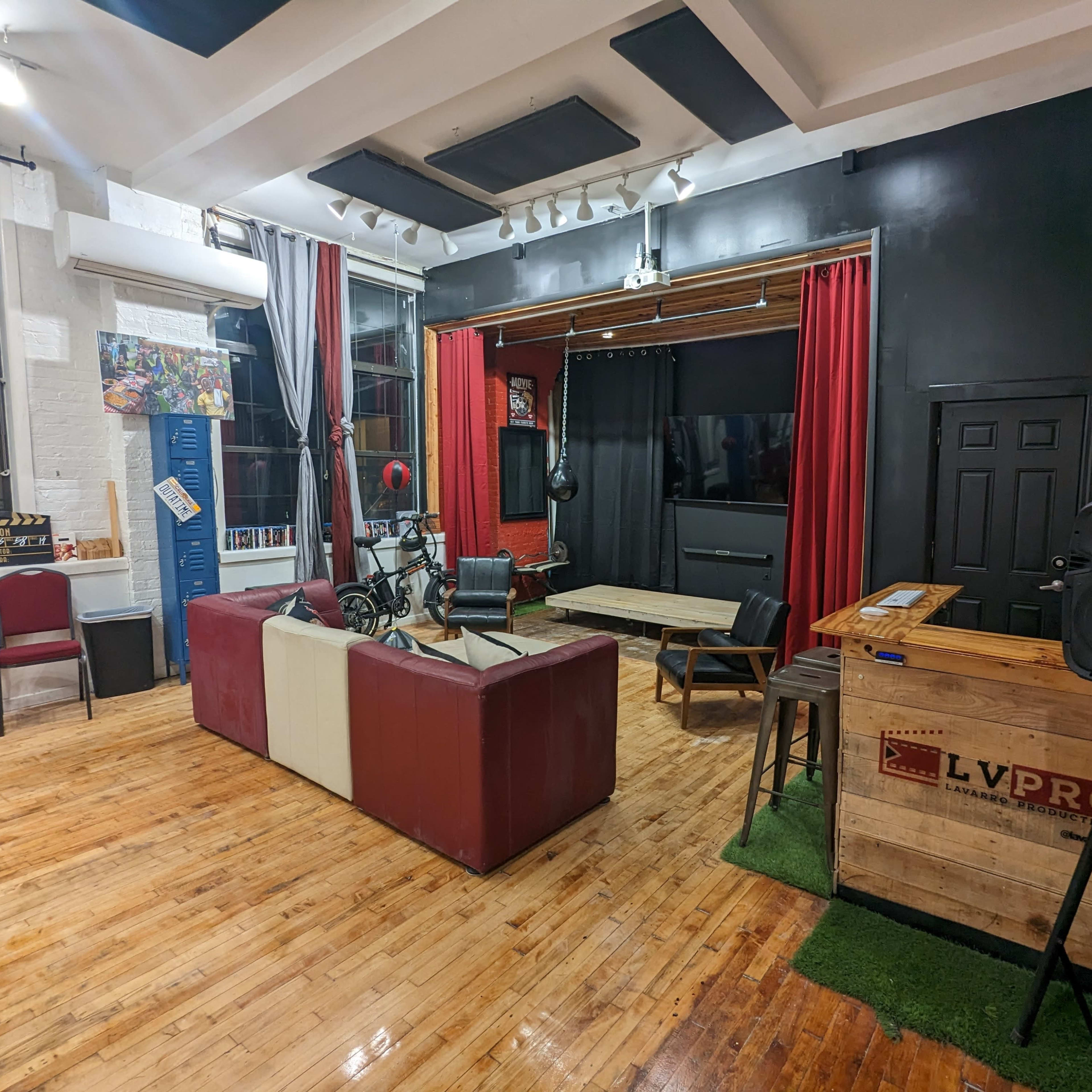 A spacious studio setting with a mix of red velvet curtains, a large screen, wooden furniture, and a combination of seating areas, including a couch and chairs.