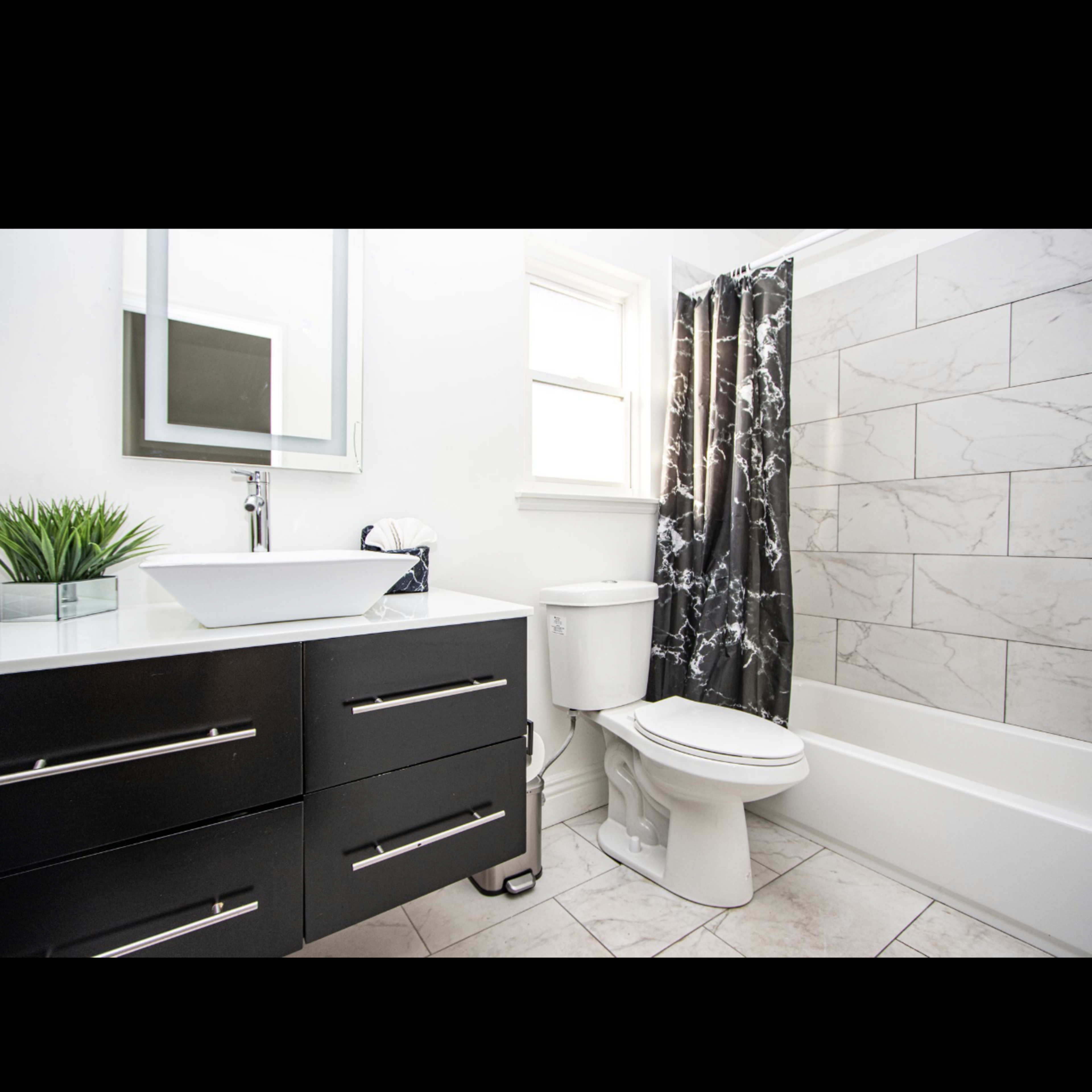 A modern bathroom features a white bathtub, a toilet, a dark vanity with drawers, and a decorative shower curtain.
