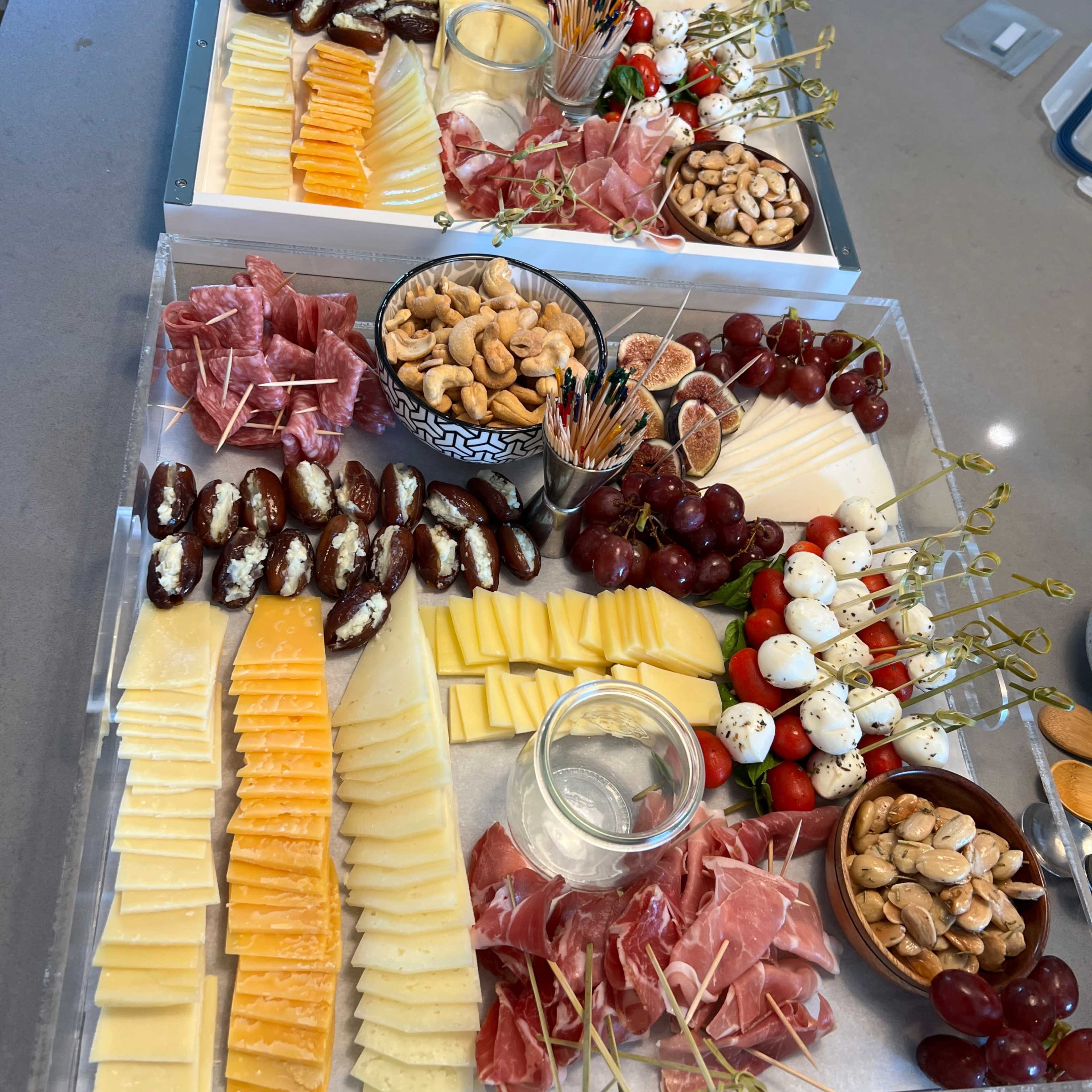 A large cheese and charcuterie board features various cheeses, cured meats, nuts, fruits, and olives arranged neatly on a rectangular tray.