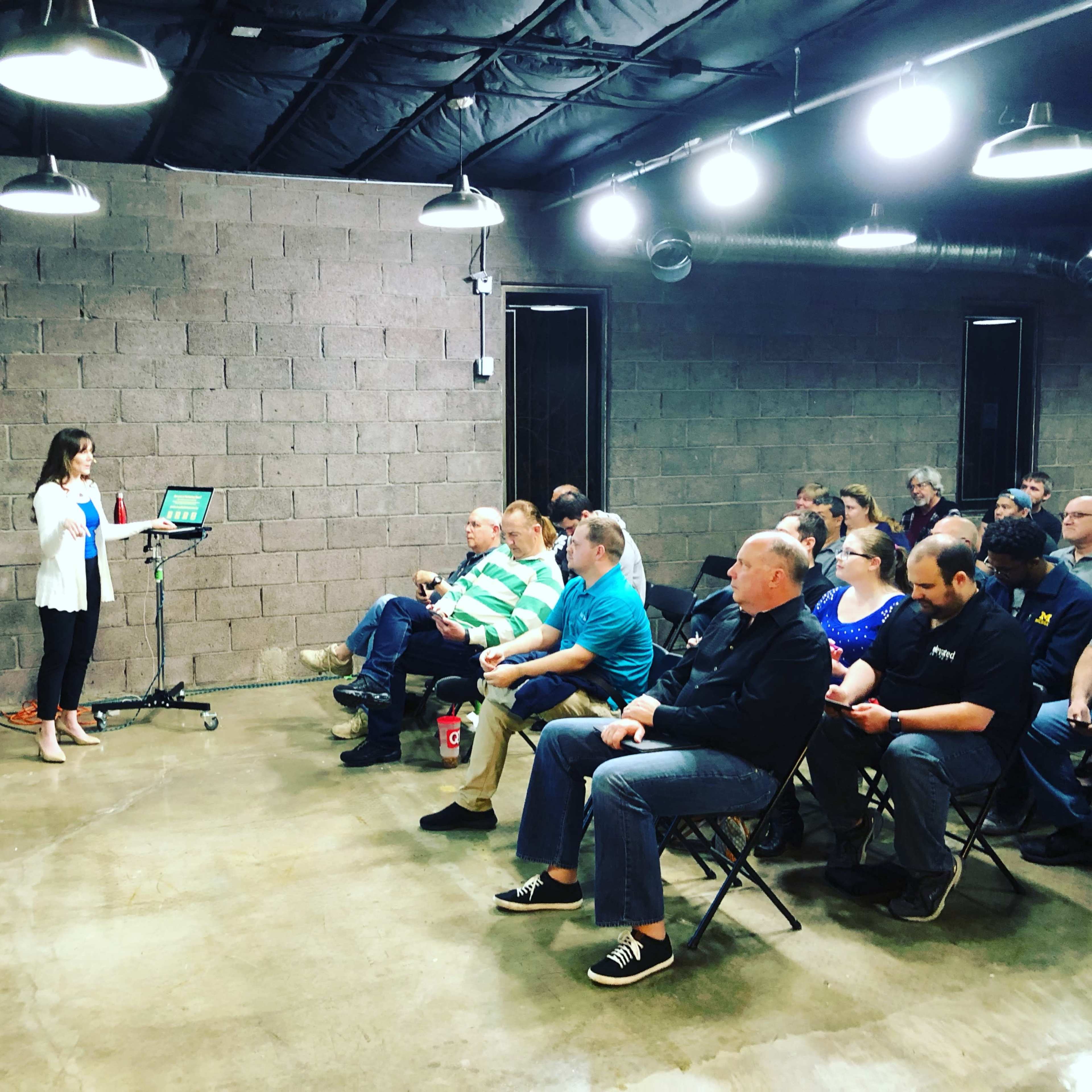 A presenter stands at a podium in front of an audience seated in folding chairs in an industrial-style meeting space.