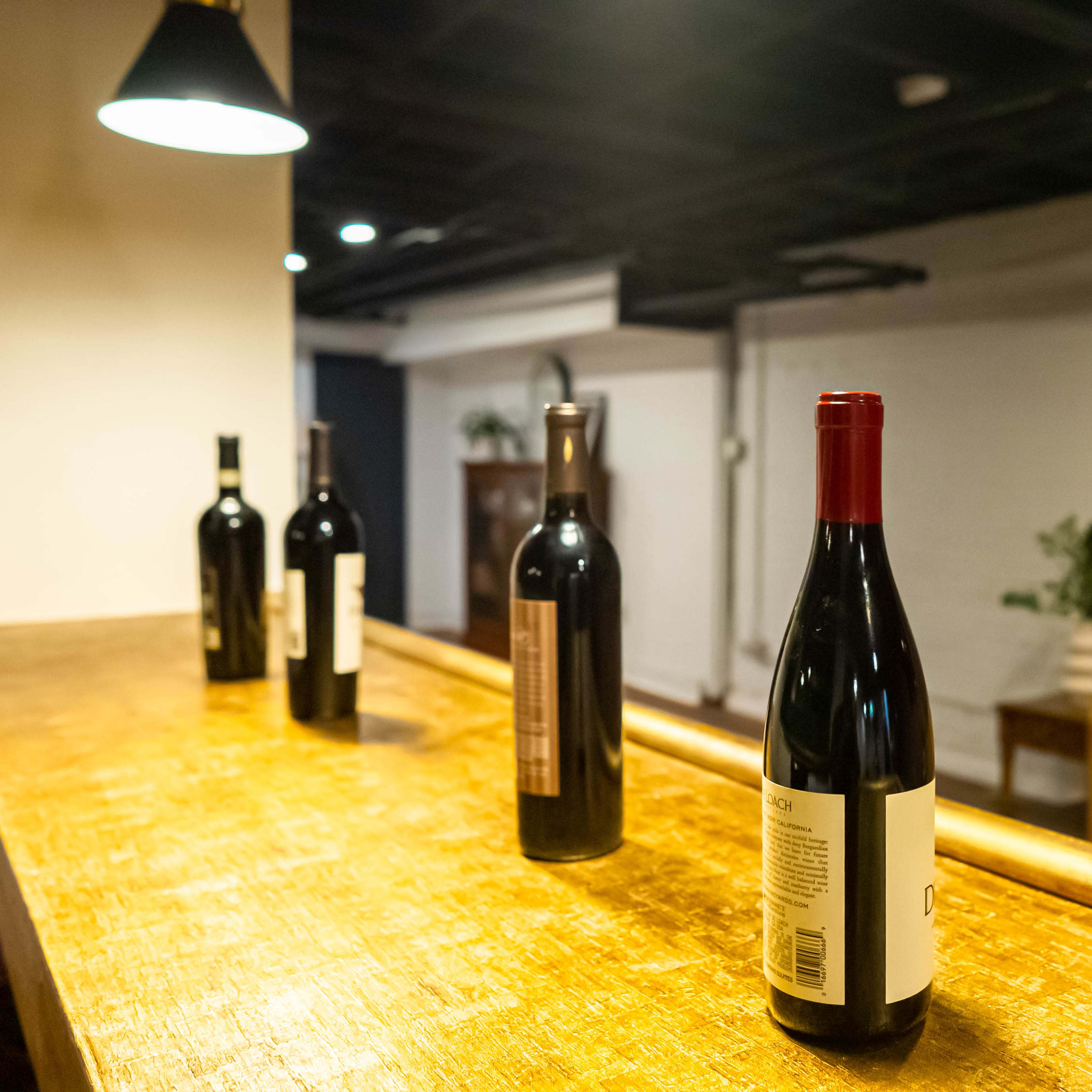 A row of wine bottles stands on a wooden surface in a dimly lit room with a yellow sofa in the background.
