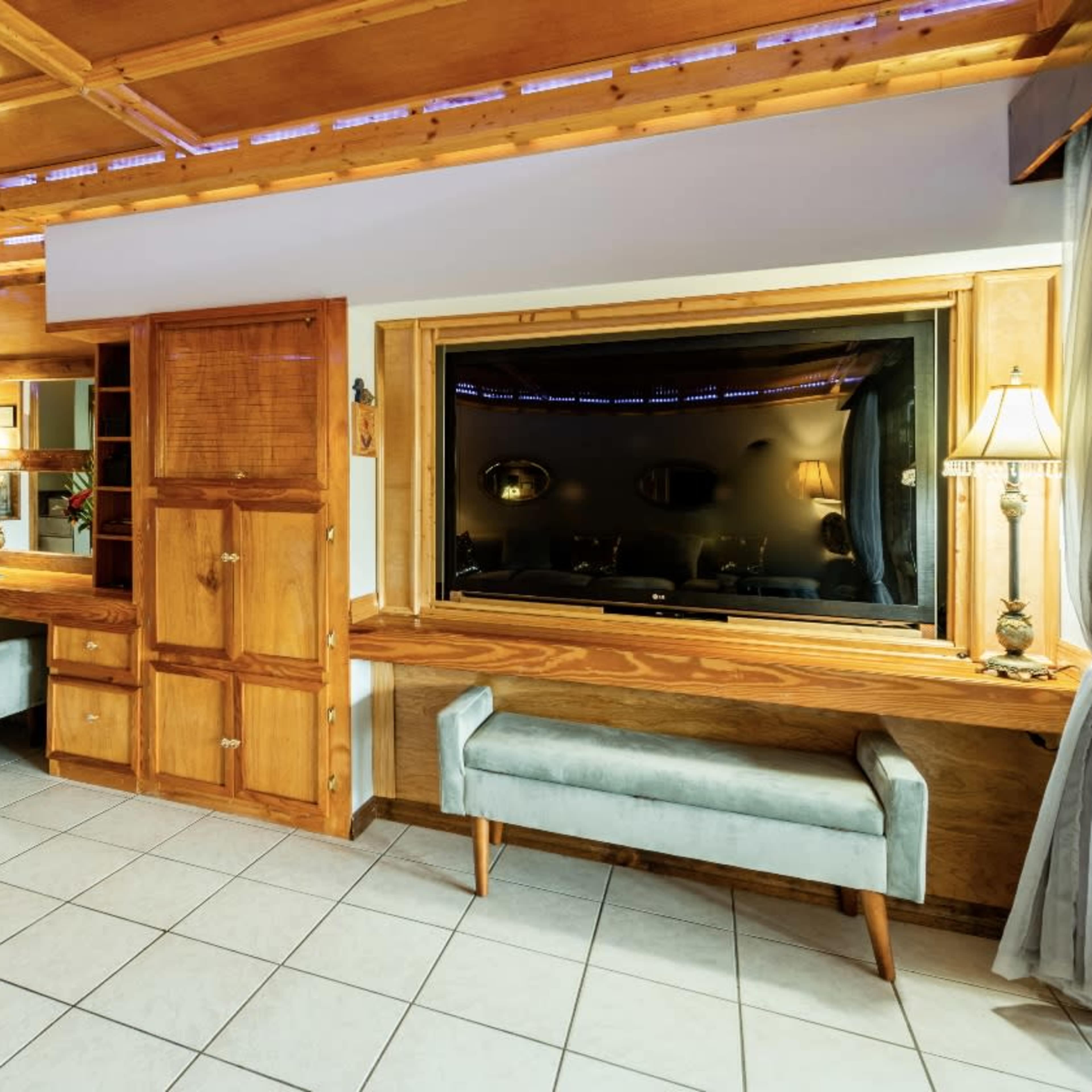A room with wooden furniture, a desk with two upholstered stools, and a large window featuring a television.