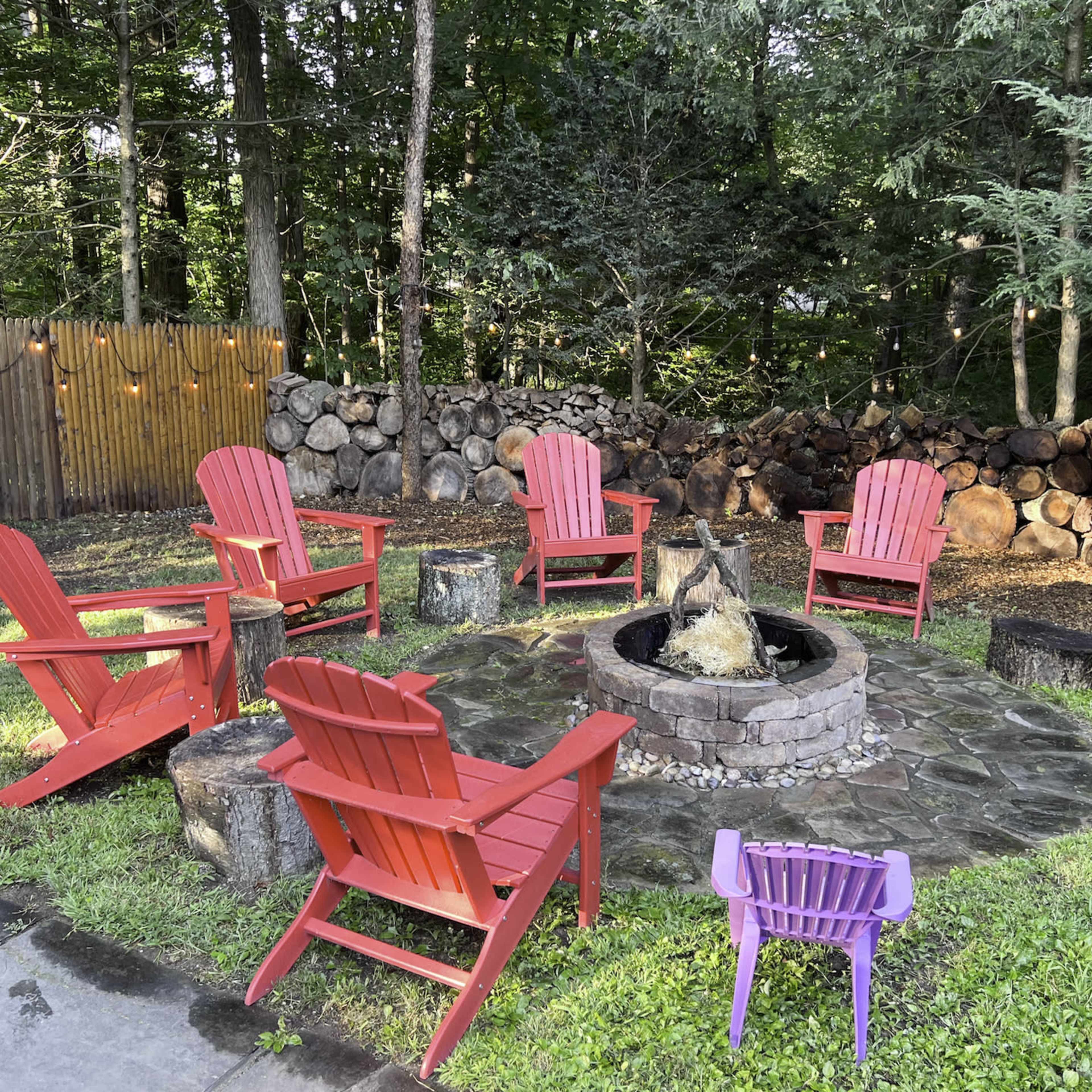 A circular seating area with six red Adirondack chairs surrounds a stone fire pit, set on a grassy lawn near a stacked woodpile and trees.
