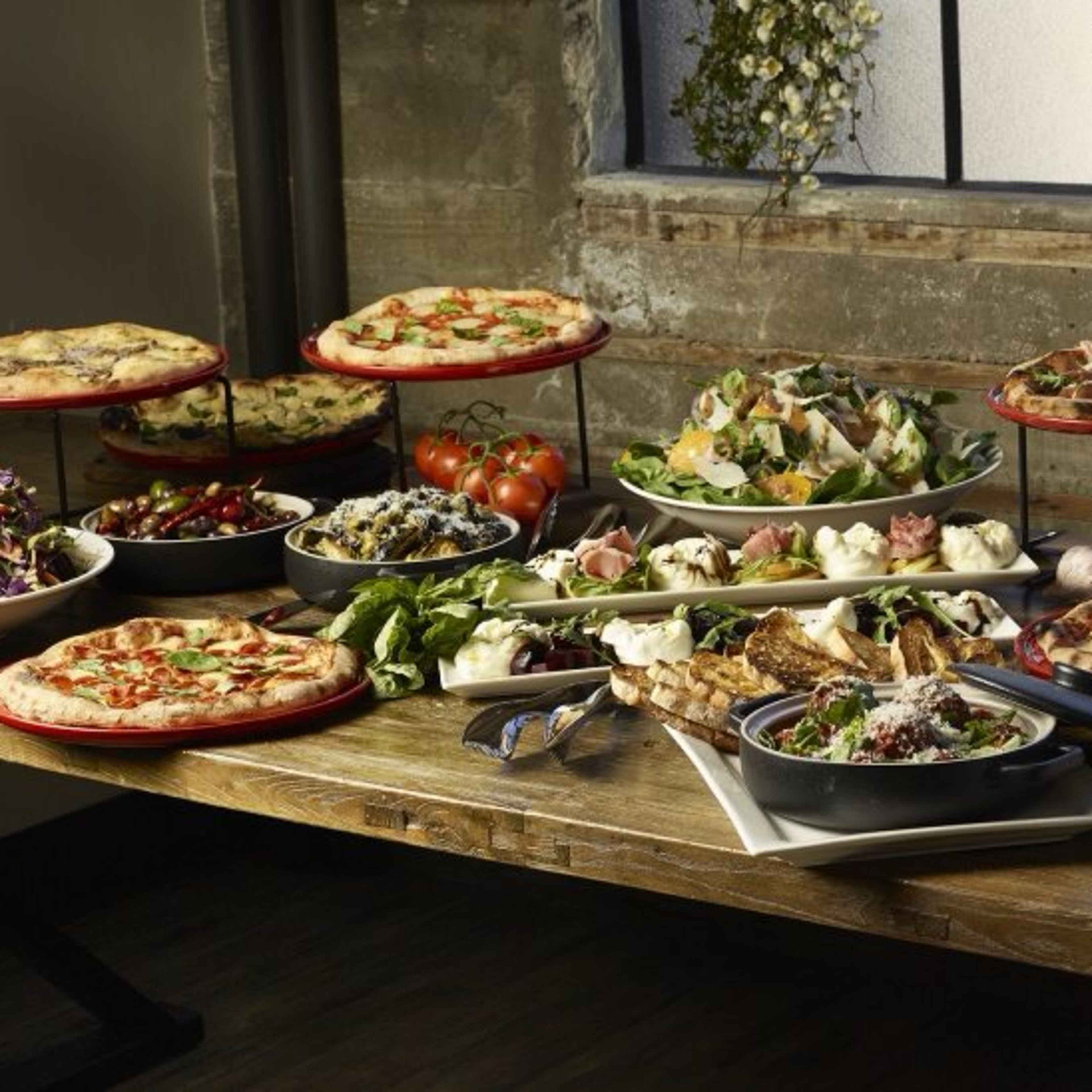 A variety of dishes, including pizzas, salads, and appetizers, are arranged on a wooden table near a large window, surrounded by potted plants.
