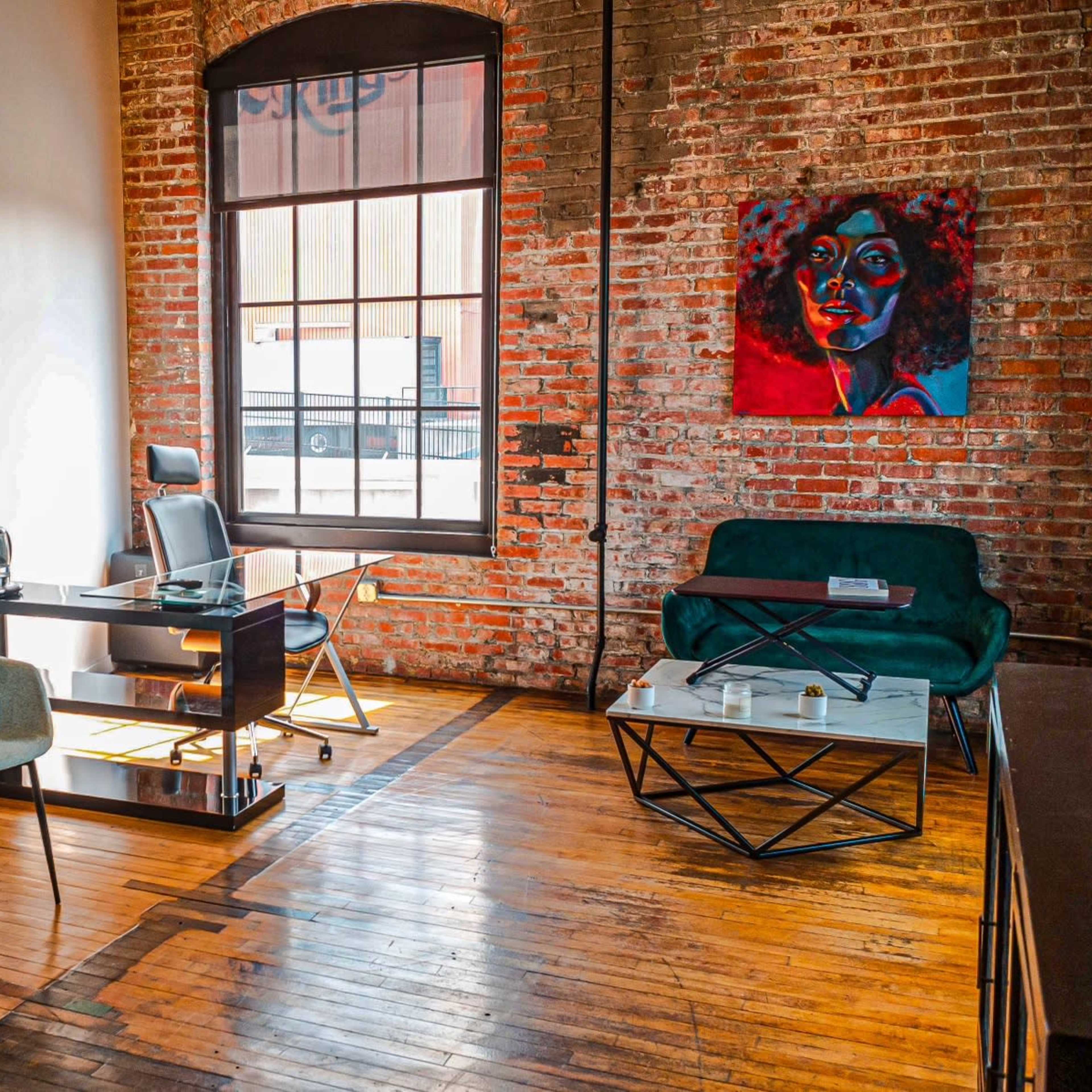 A stylish office space featuring a wooden floor, a brick wall, a desk with a chair, two armchairs, and a coffee table, along with a colorful painting on the wall.