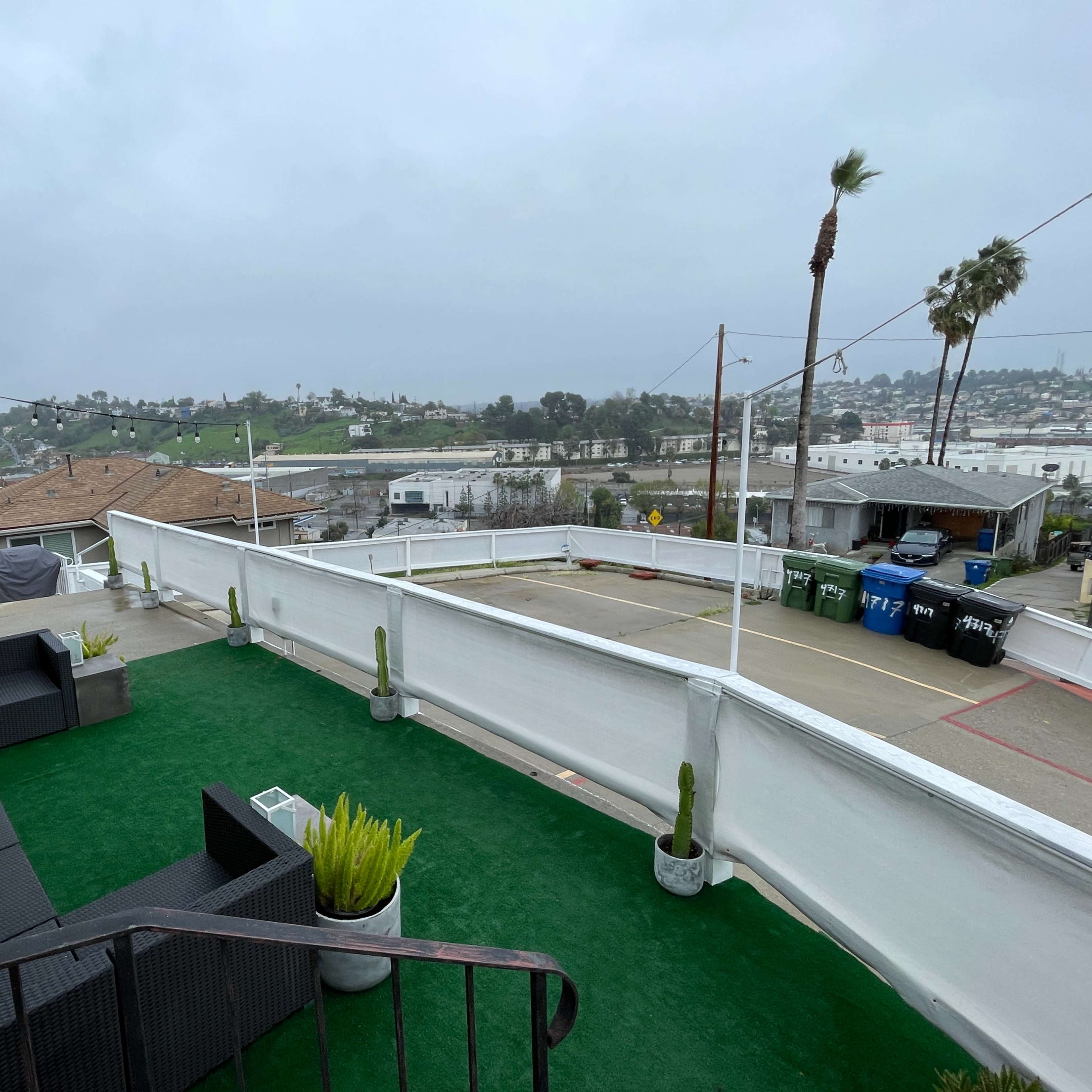 A rooftop terrace with green artificial grass, lounging furniture, and a view of hills and buildings in the background under overcast skies.