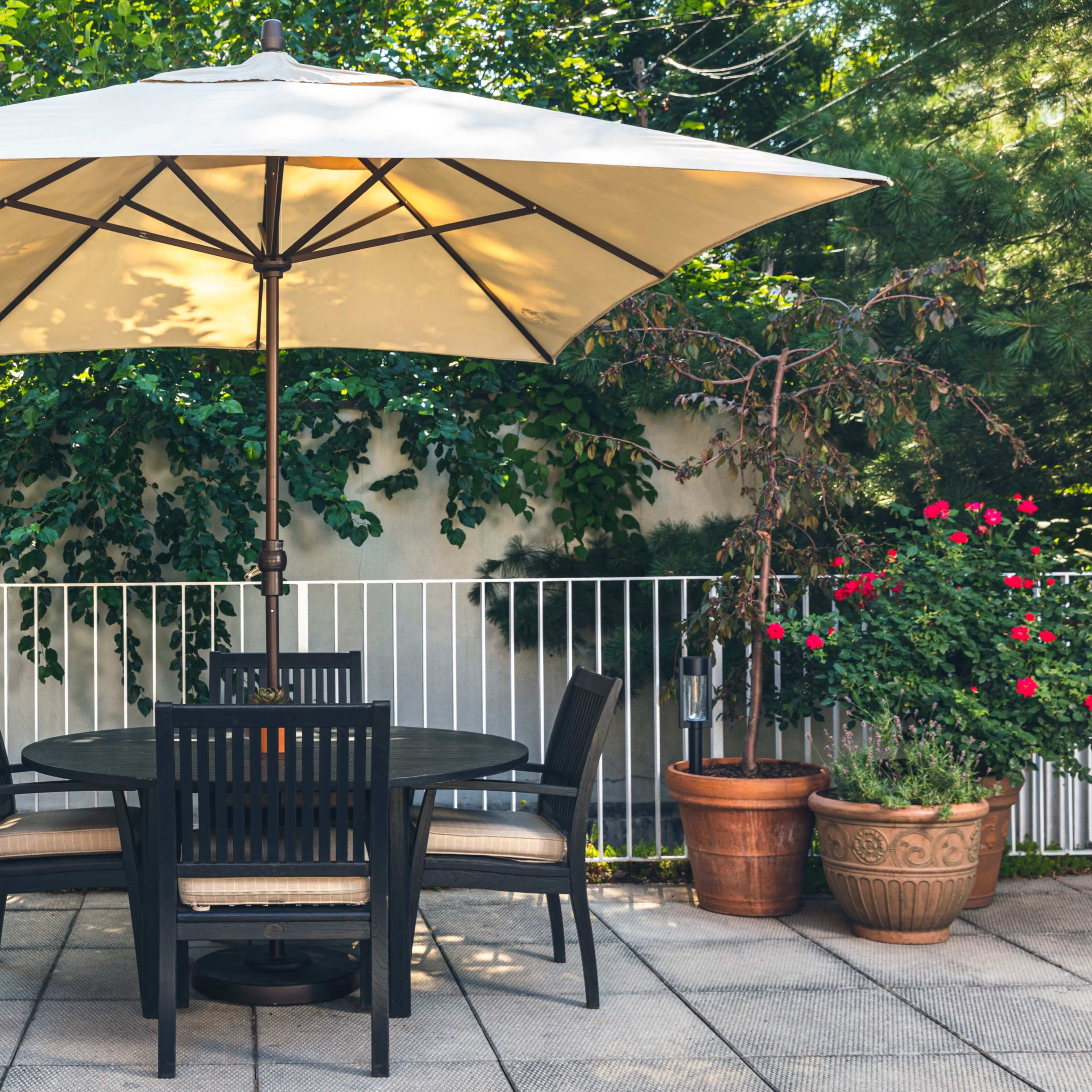 A circular table with four chairs sits under a large umbrella on a patio surrounded by potted plants and a white fence.