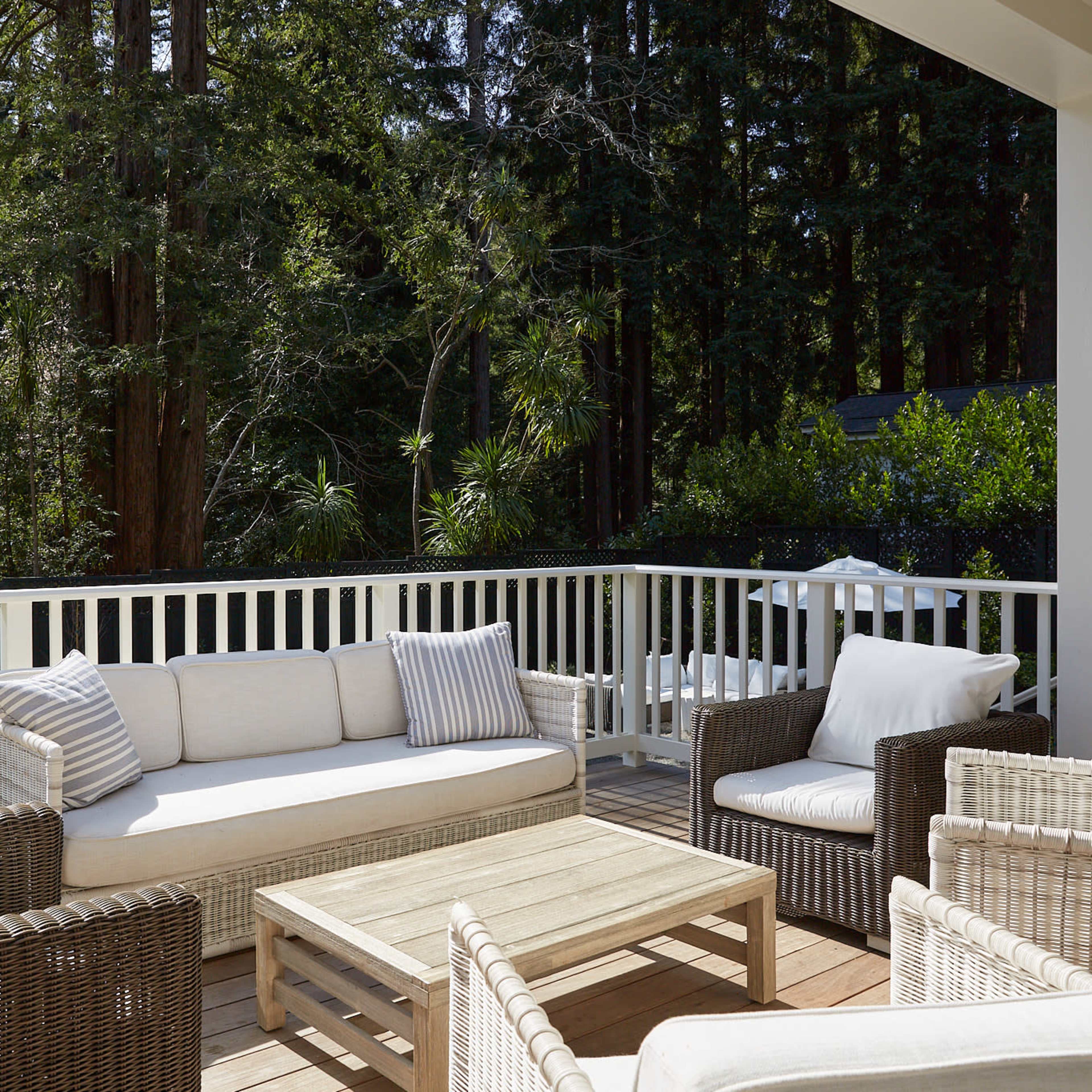 A modern patio features a white sofa and wicker chairs arranged around a wooden coffee table, surrounded by lush greenery.
