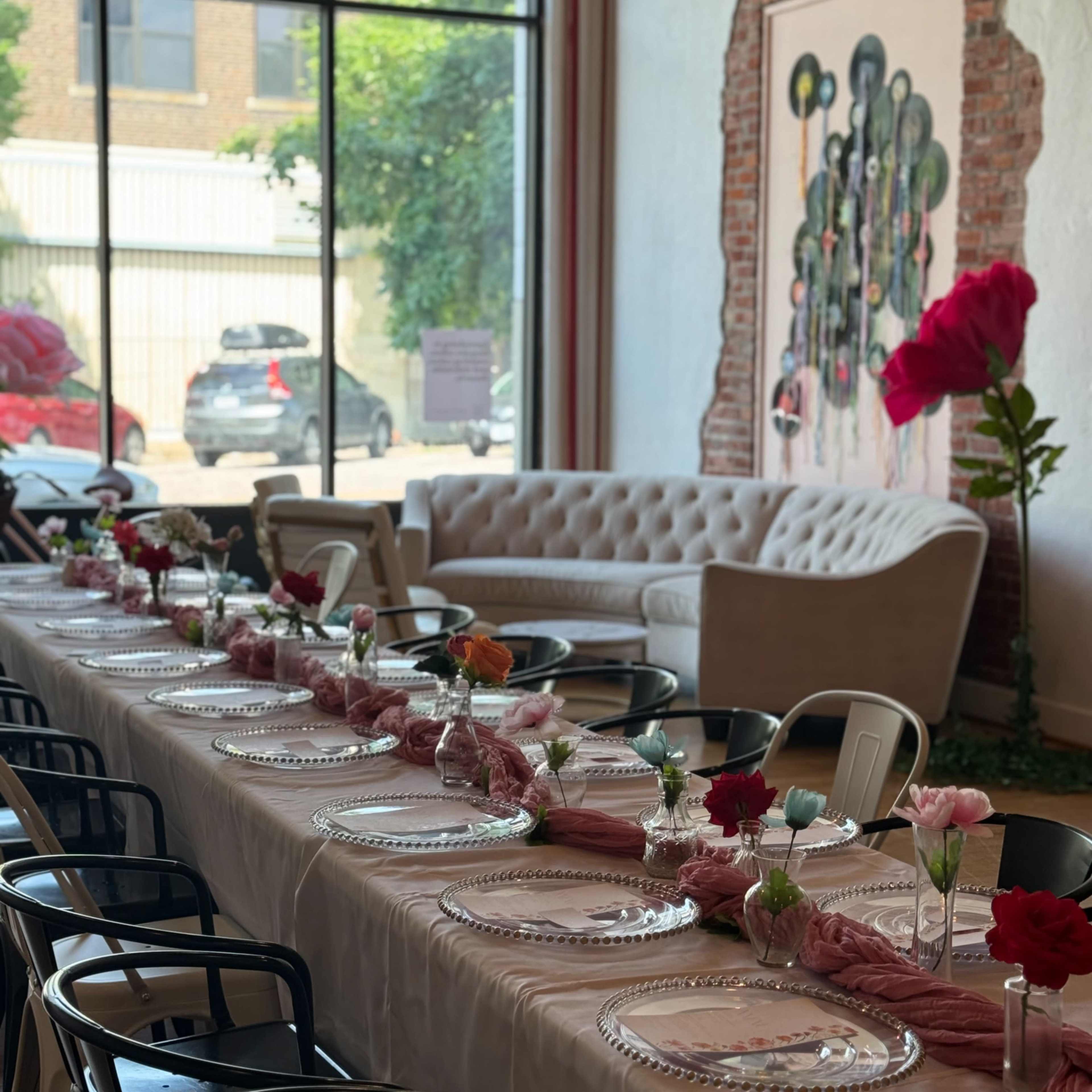 A long dining table is set with plates and flowers in a bright room featuring large windows and a couch.