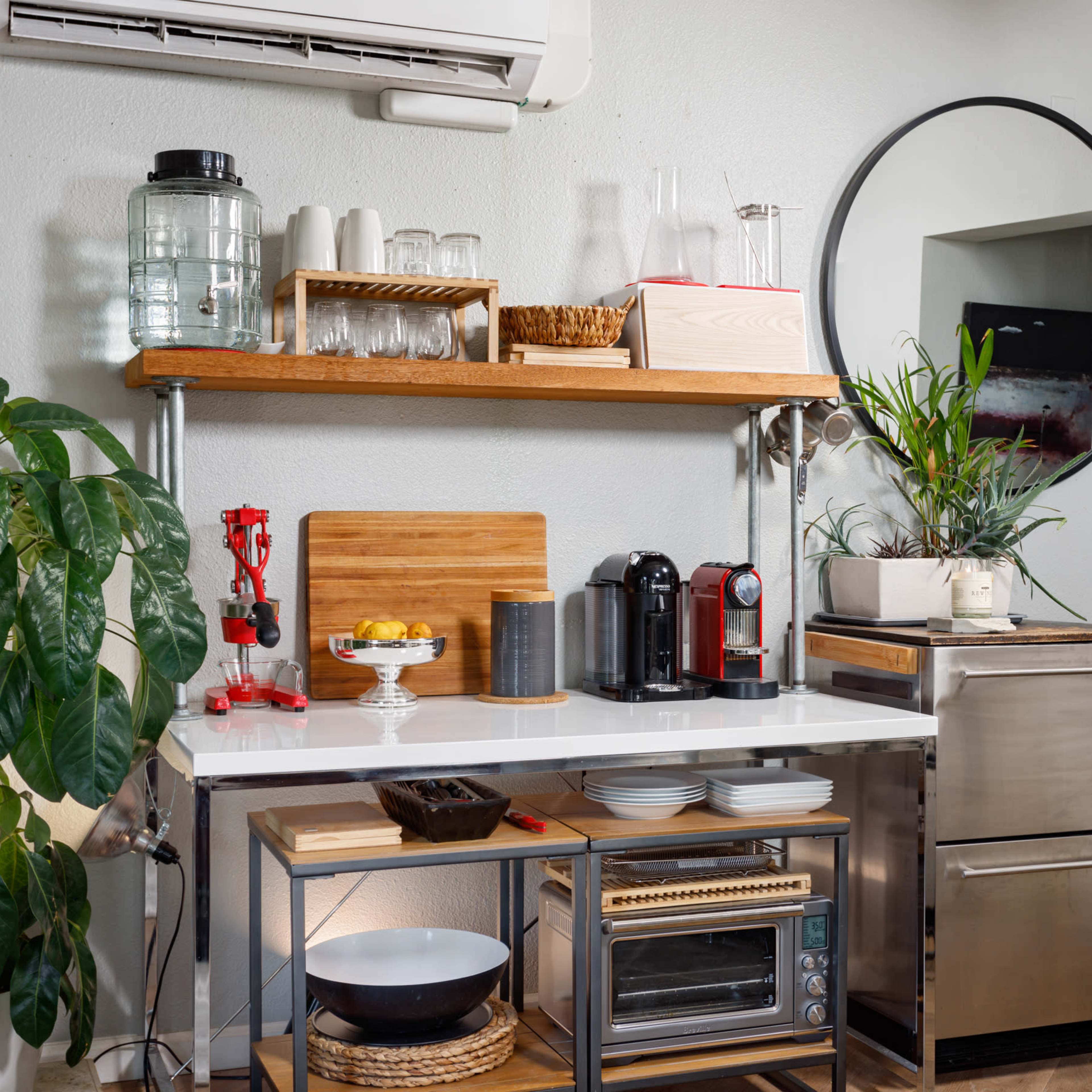 A modern kitchen area featuring a countertop with a coffee machine, fruits, and utensils, along with shelves holding glassware and a large mirror.