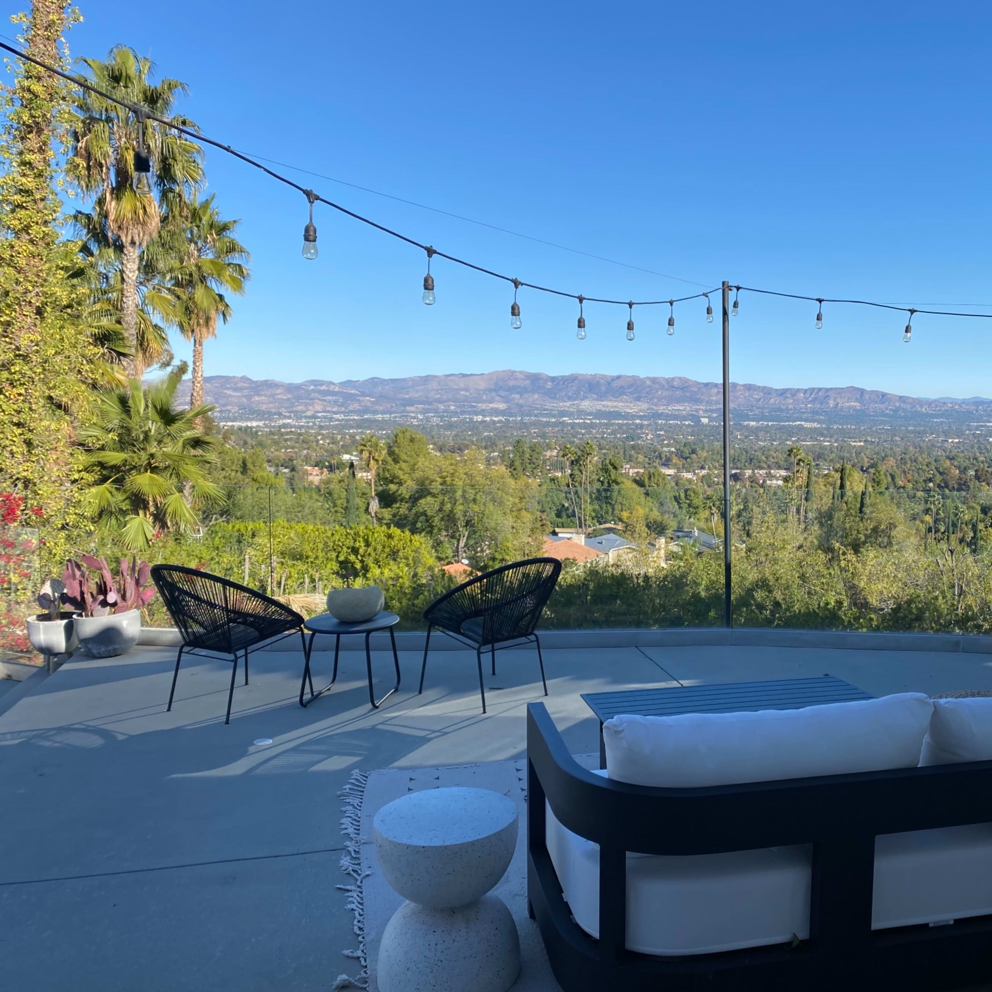 A patio with modern seating overlooks a valley and mountains under a clear blue sky, illuminated by string lights.