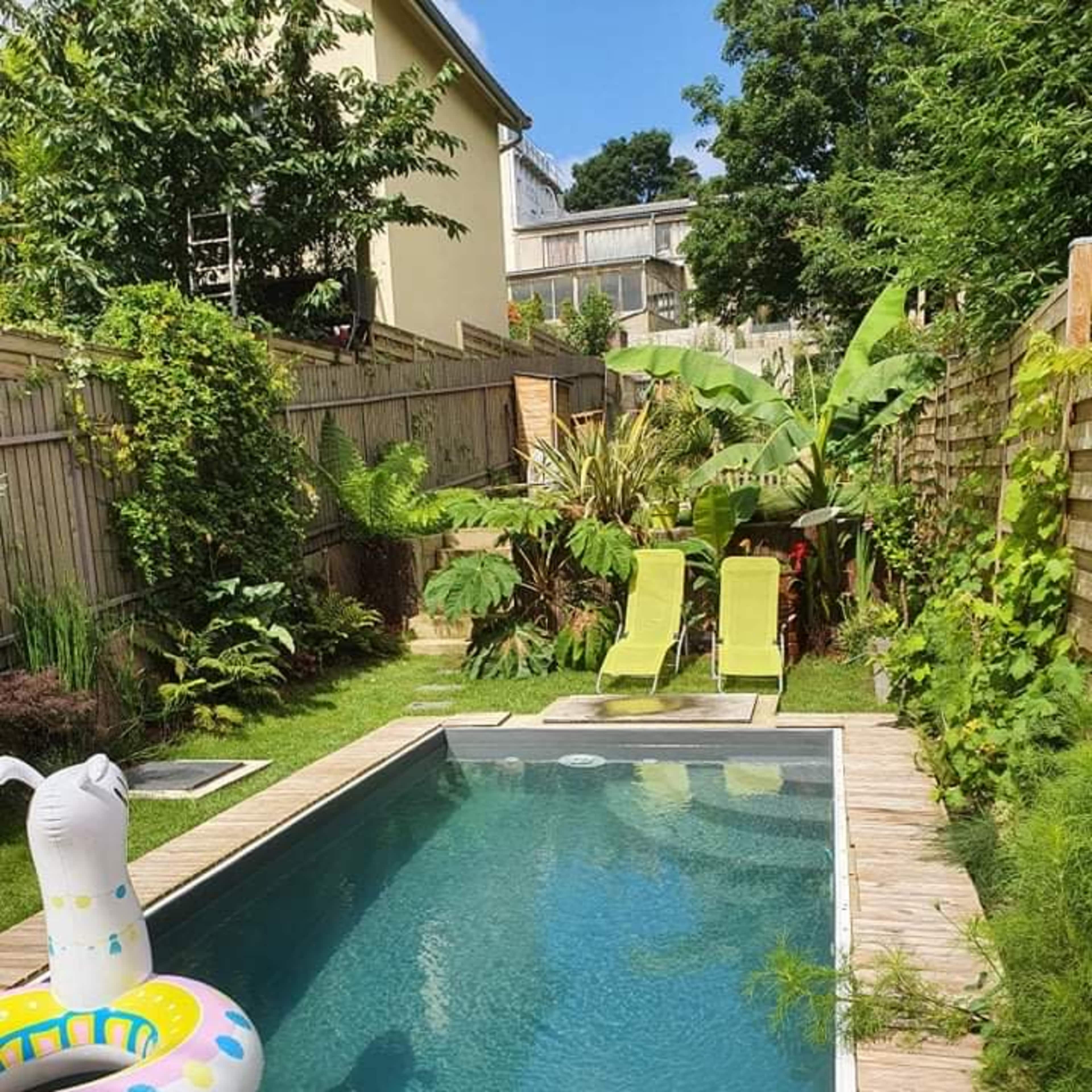 A backyard pool surrounded by greenery, featuring two lounge chairs and a colorful inflatable float in the water.