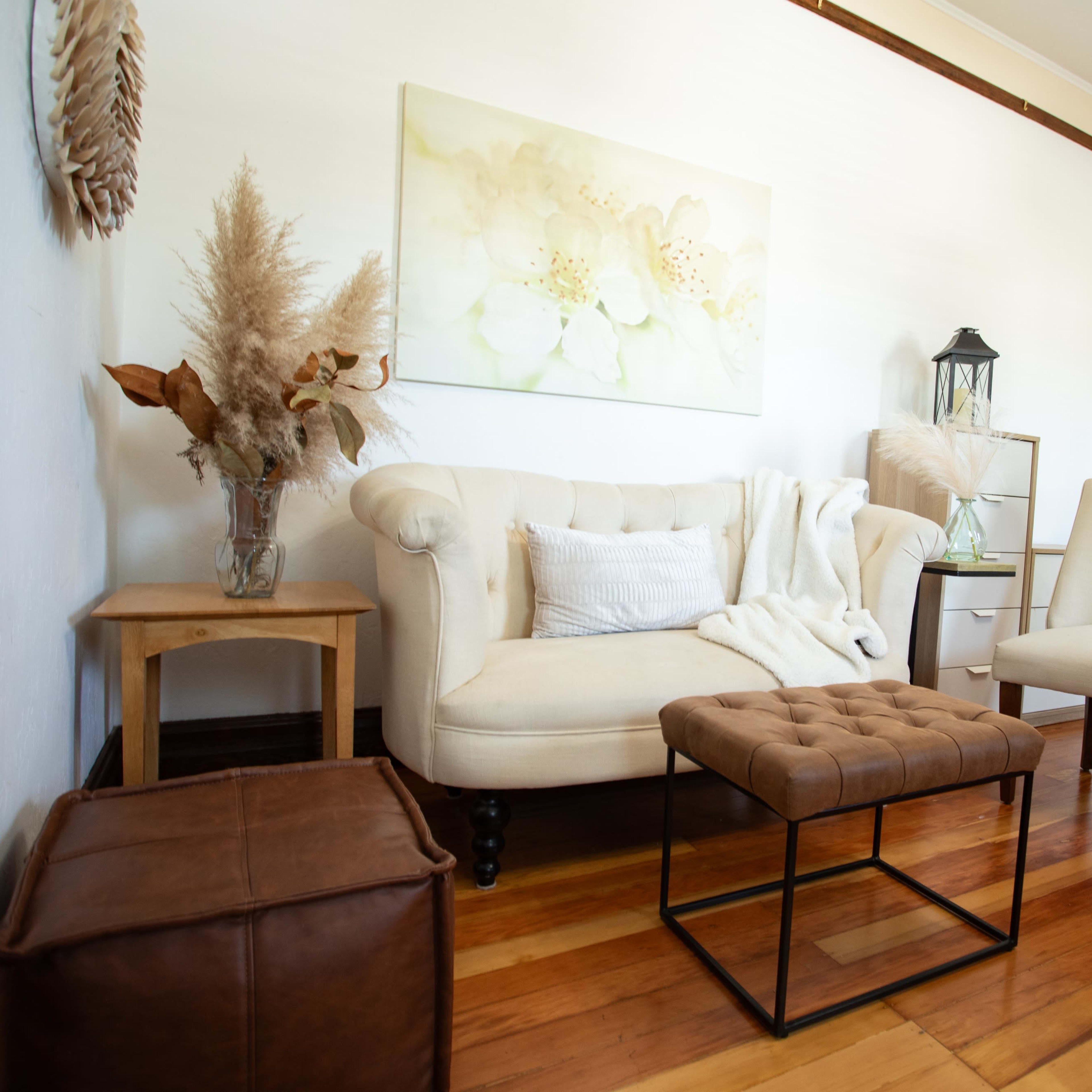 A cozy living room with a beige tufted sofa, a small wooden side table, a decorative armchair, and a brown leather ottoman, all situated on polished wooden floors.