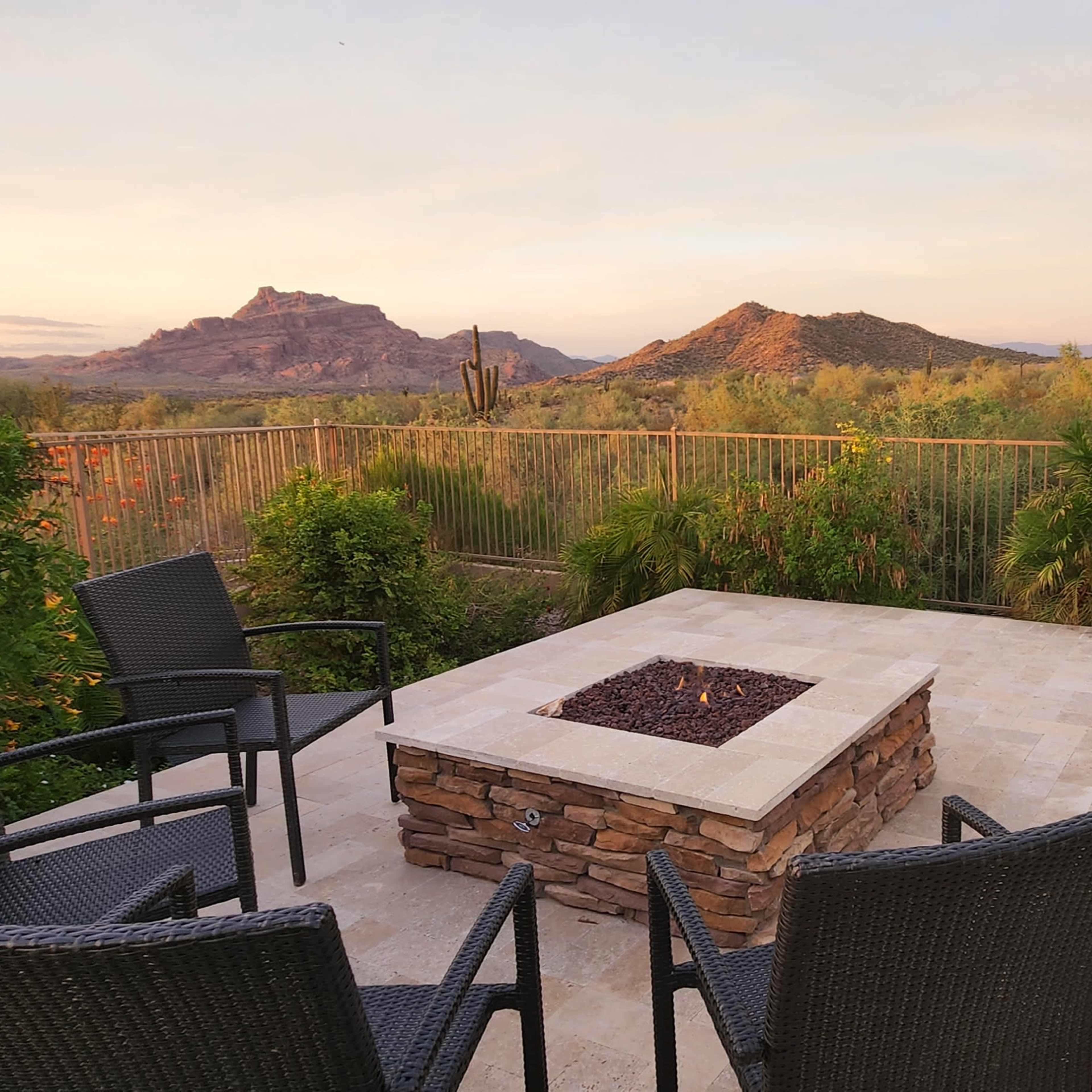 A patio area with black chairs facing a stone fire pit and a small pool, overlooking a desert landscape with mountains in the background.