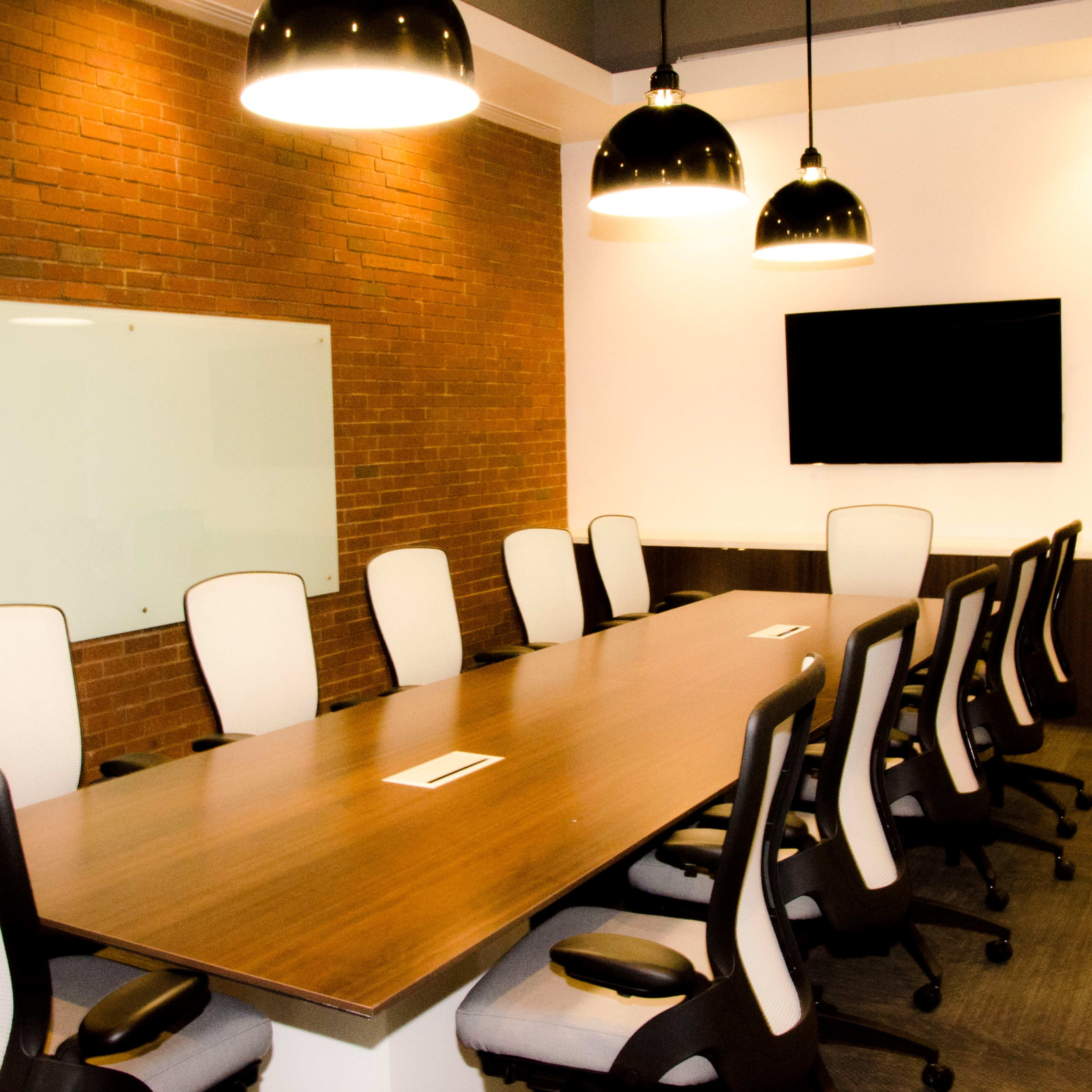 A conference room with a long wooden table surrounded by eight office chairs, a large screen on the wall, and a brick accent wall.