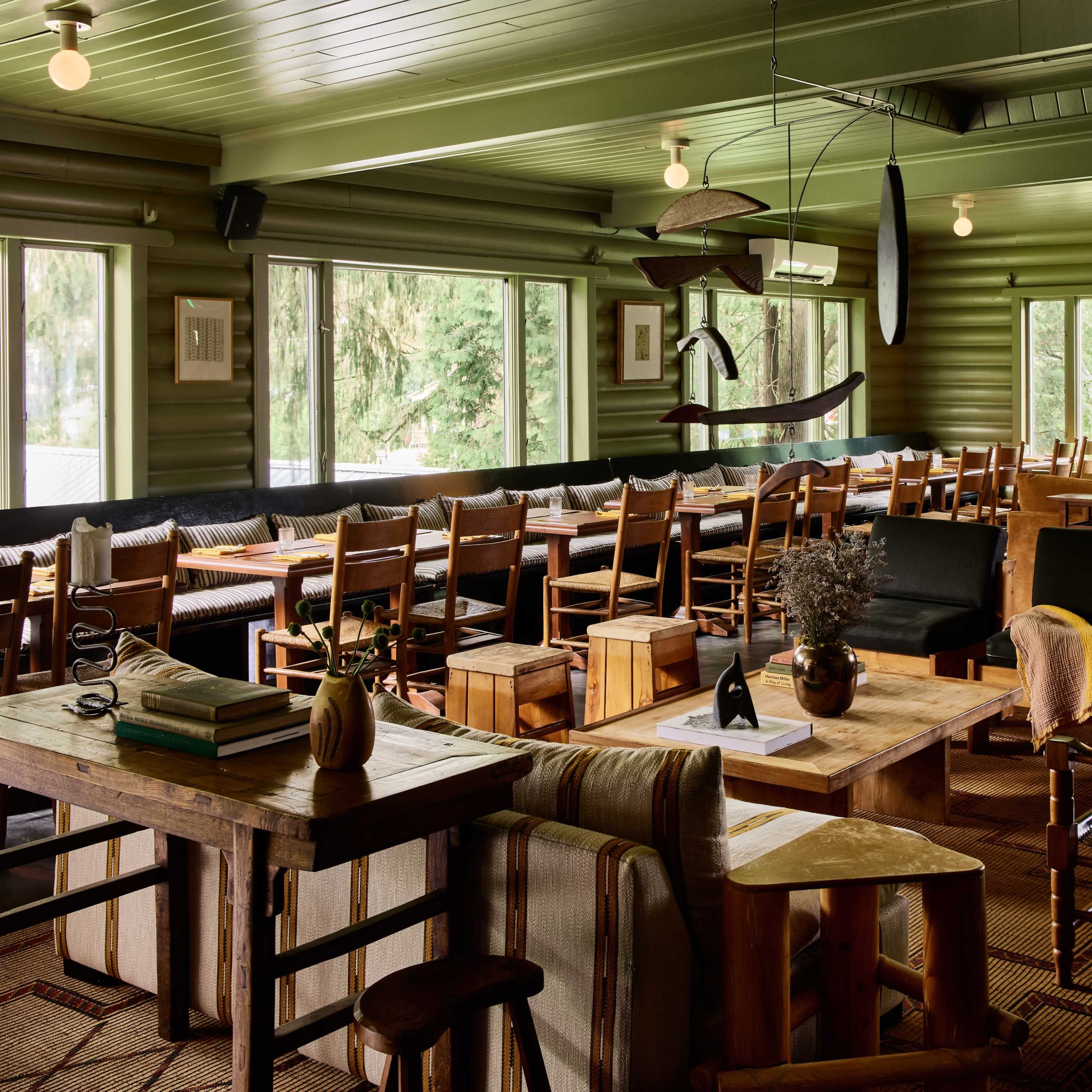 A spacious dining area with wooden tables and chairs, large windows allowing natural light, and a stone fireplace at one end.