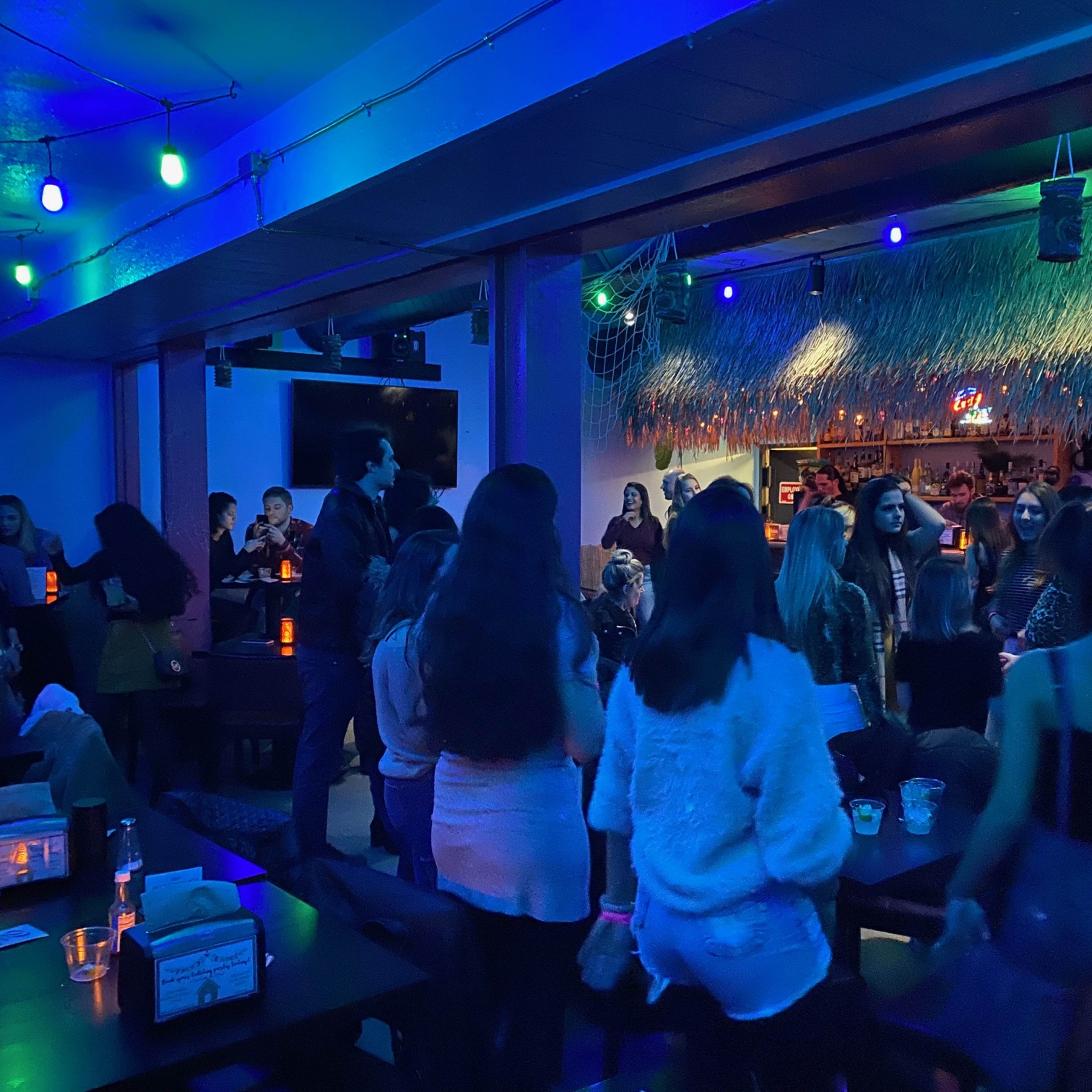 A lively crowd of people gathers in a dimly lit bar decorated with colorful lights and a thatched roof.