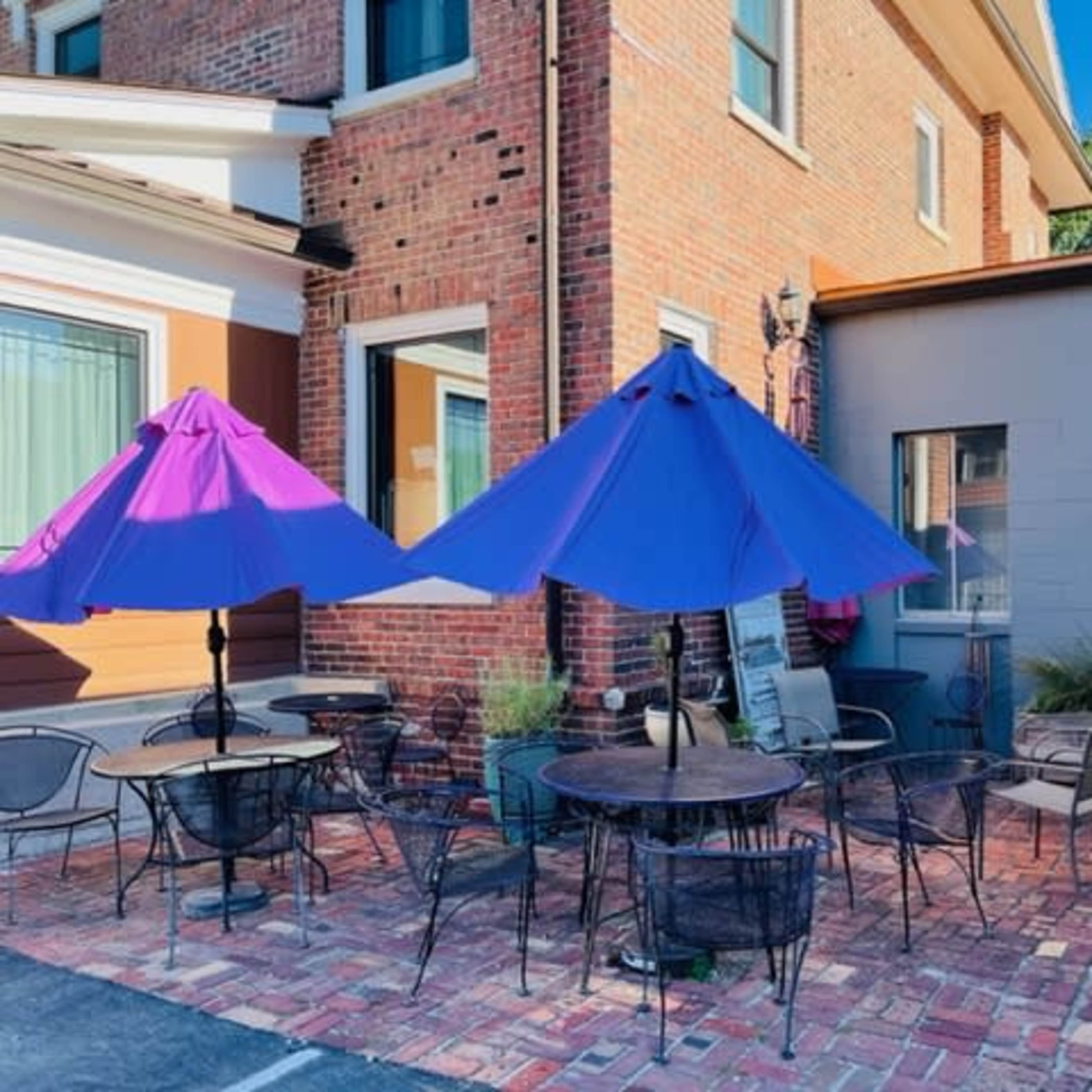 A brick-paved outdoor seating area with several round tables and purple umbrellas outside a building.