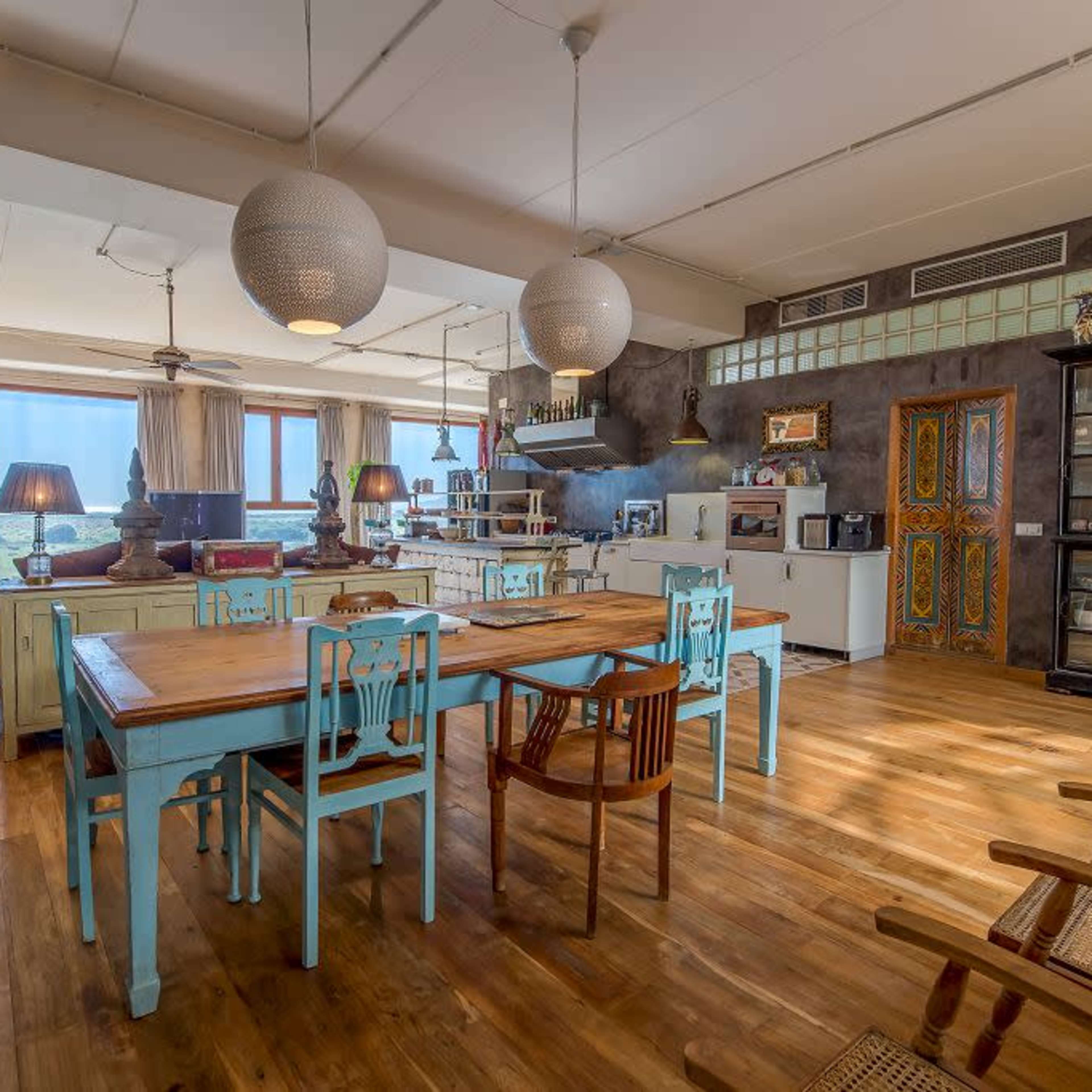 A spacious dining area with a large wooden table and colorful chairs, surrounded by shelving units and large windows letting in natural light.