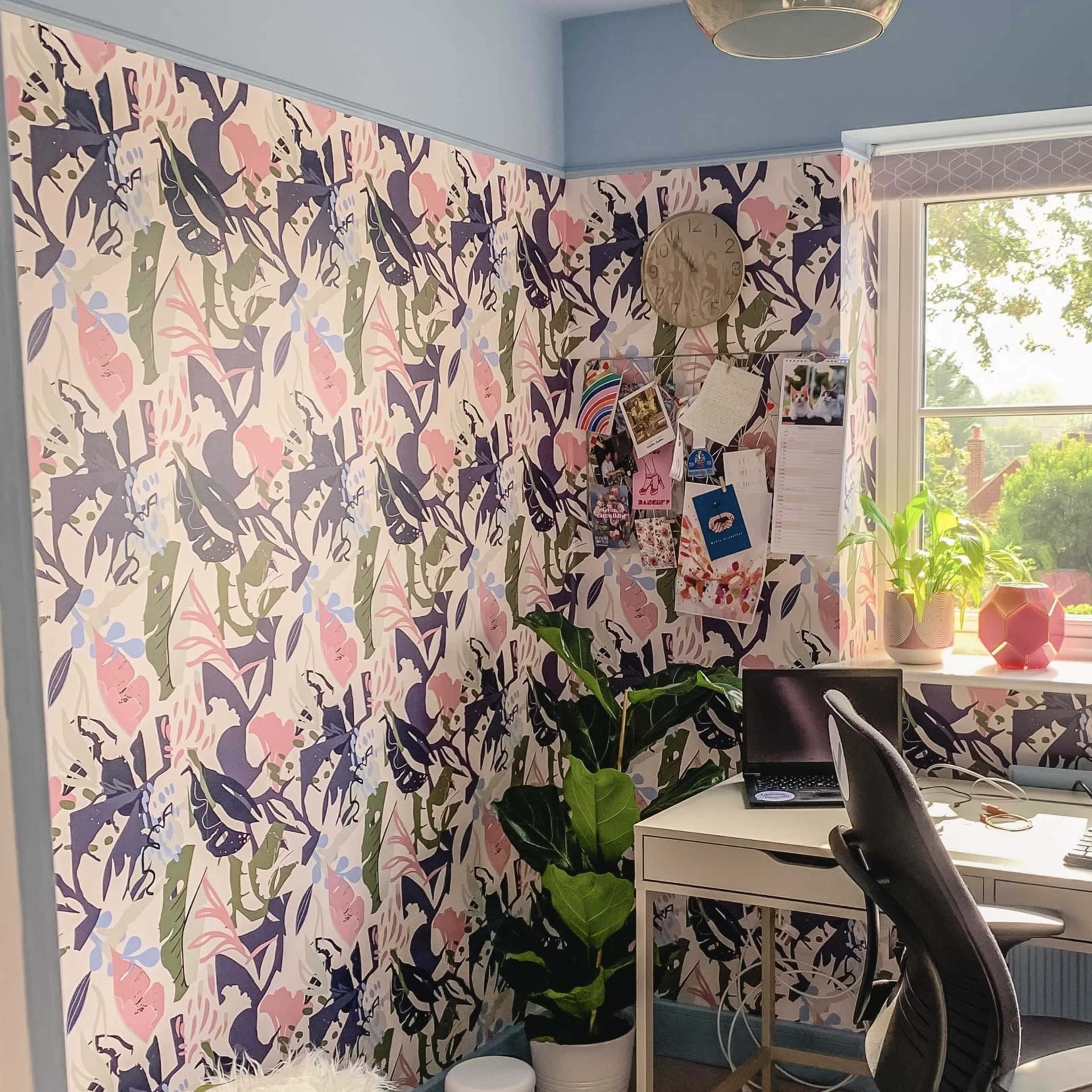 A bright home office features a desk with a computer, a chair, and decorative plants against a colorful floral wallpaper.
