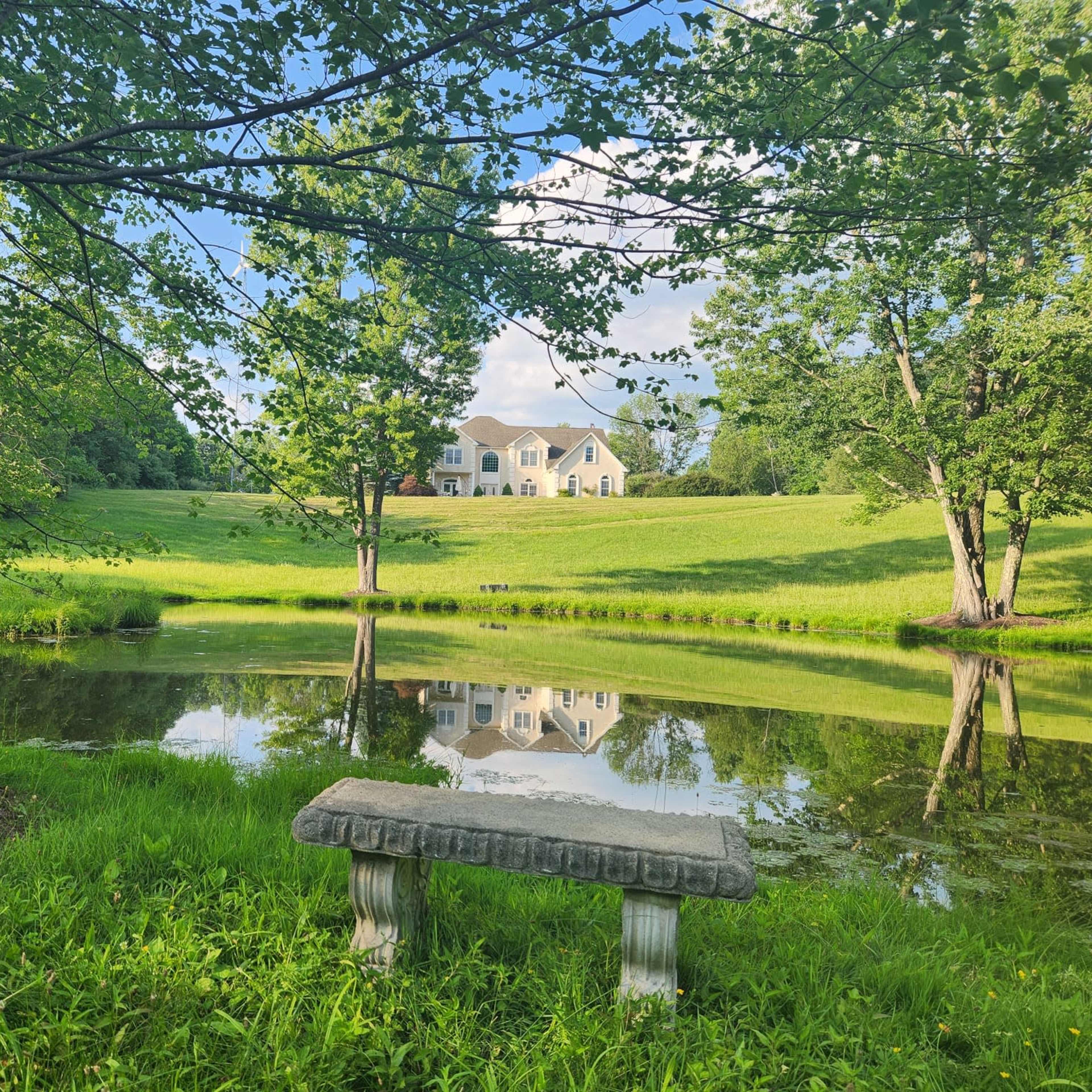 A stone bench sits beside a still pond, reflecting a large house surrounded by green trees and grassy hills.