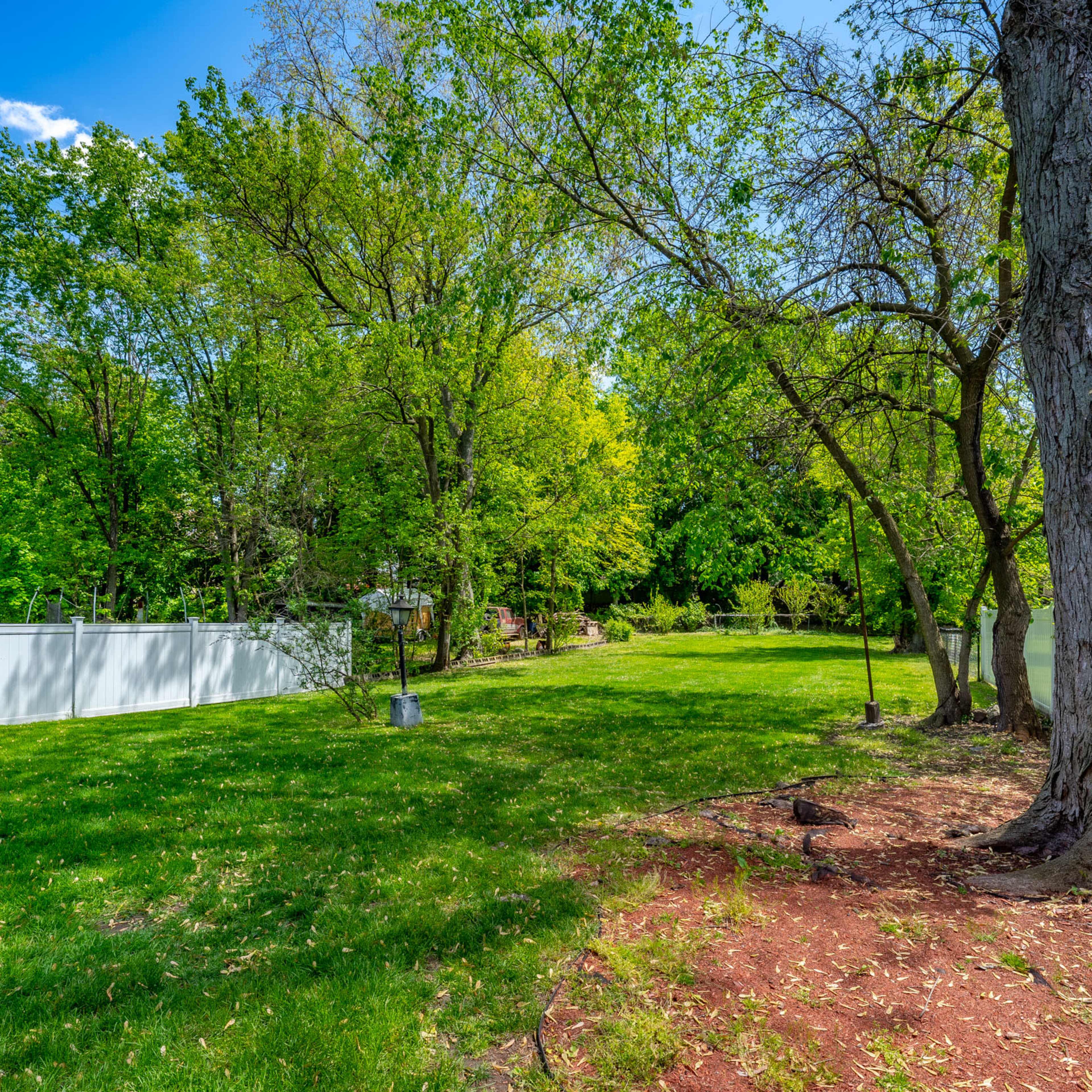 A spacious back yard with lush green grass, trees, and a white fence lining one side.