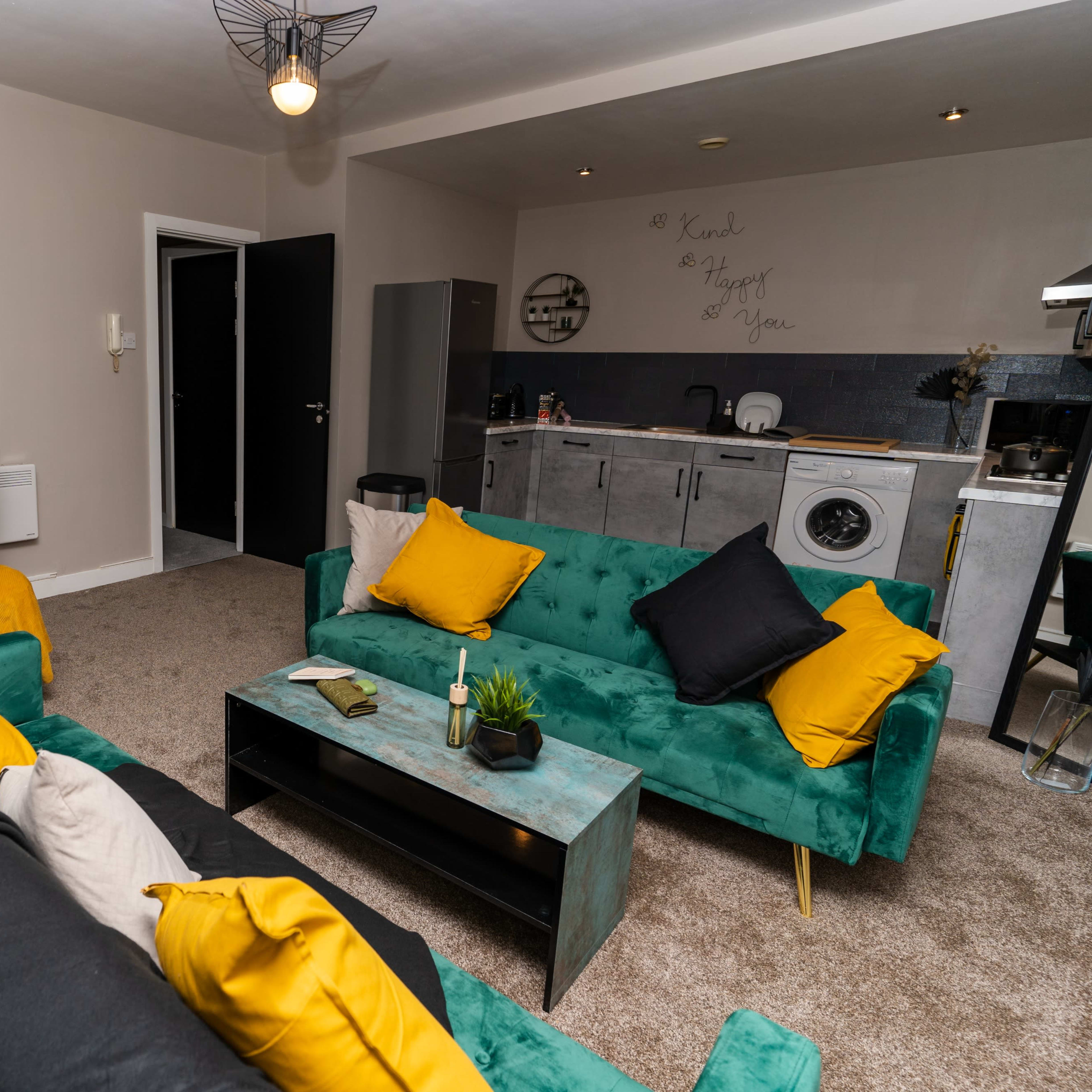 A modern living area featuring a seating arrangement with green and black sofas, a dining table set for four, and a kitchen area in the background.