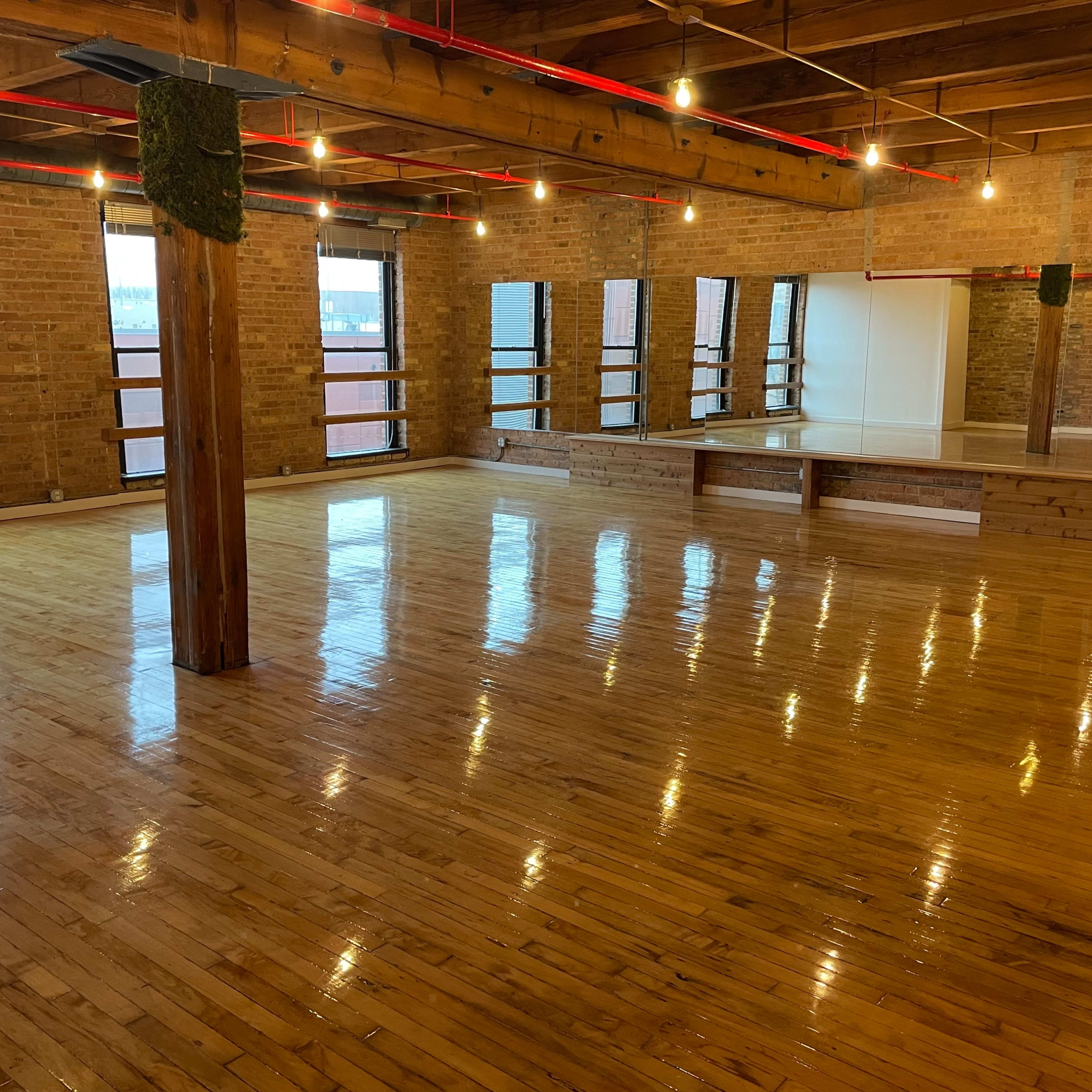 A spacious room with polished wooden floors, exposed brick walls, and large windows allowing natural light to enter.