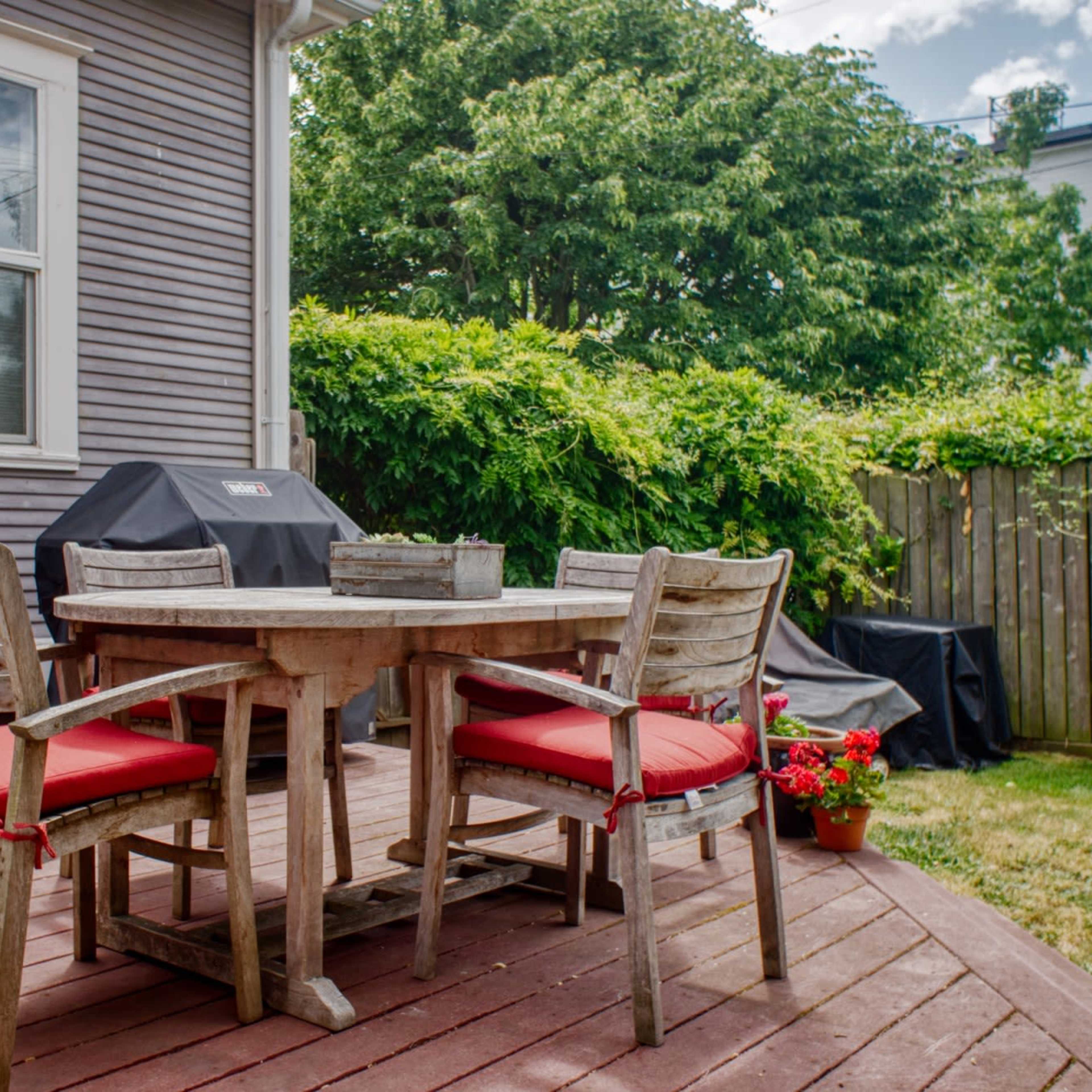 A wooden outdoor dining table with red cushions sits on a deck surrounded by greenery and a grill in the background.