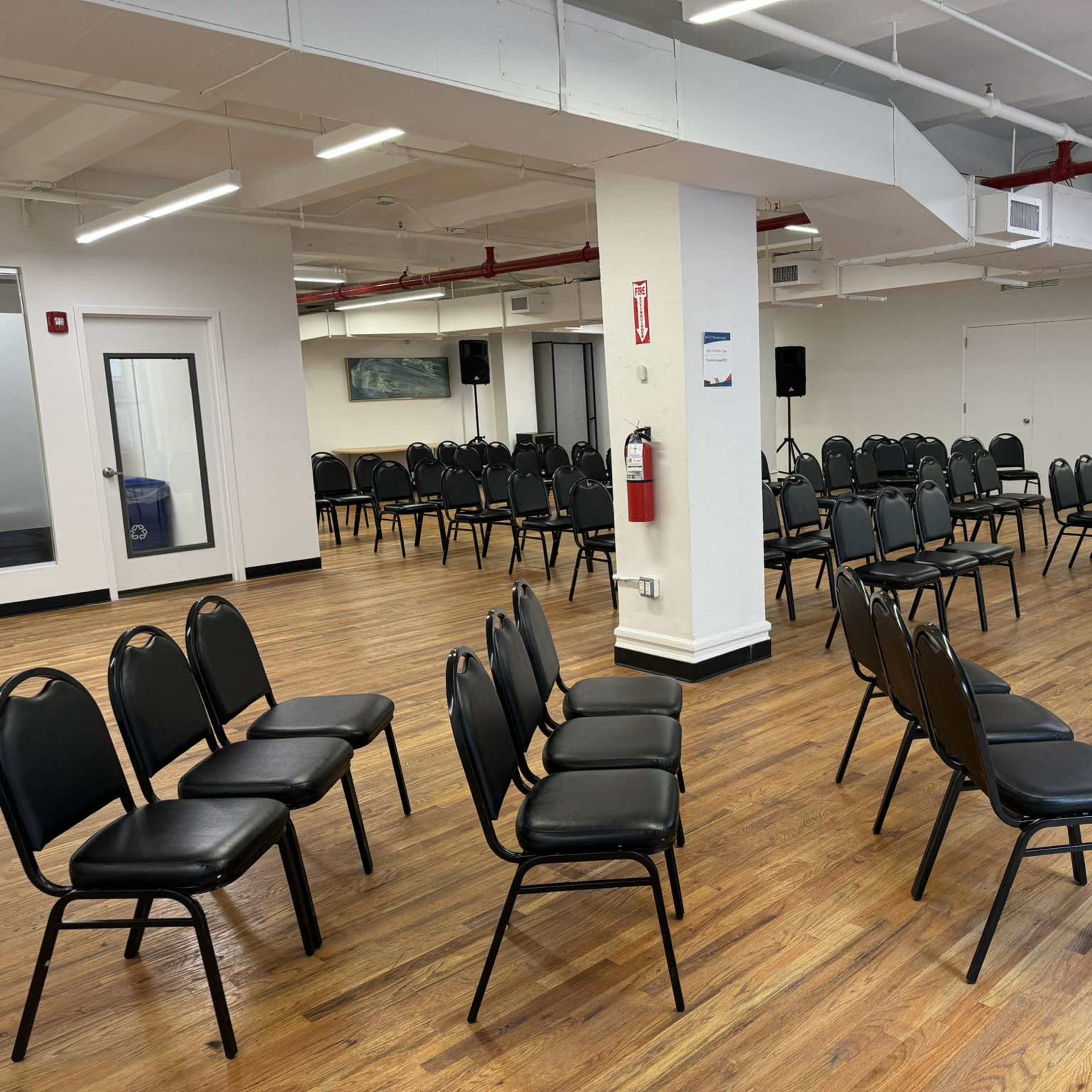 A spacious room with several rows of black chairs arranged in a semi-circle, and a single white table with a chair at one end.