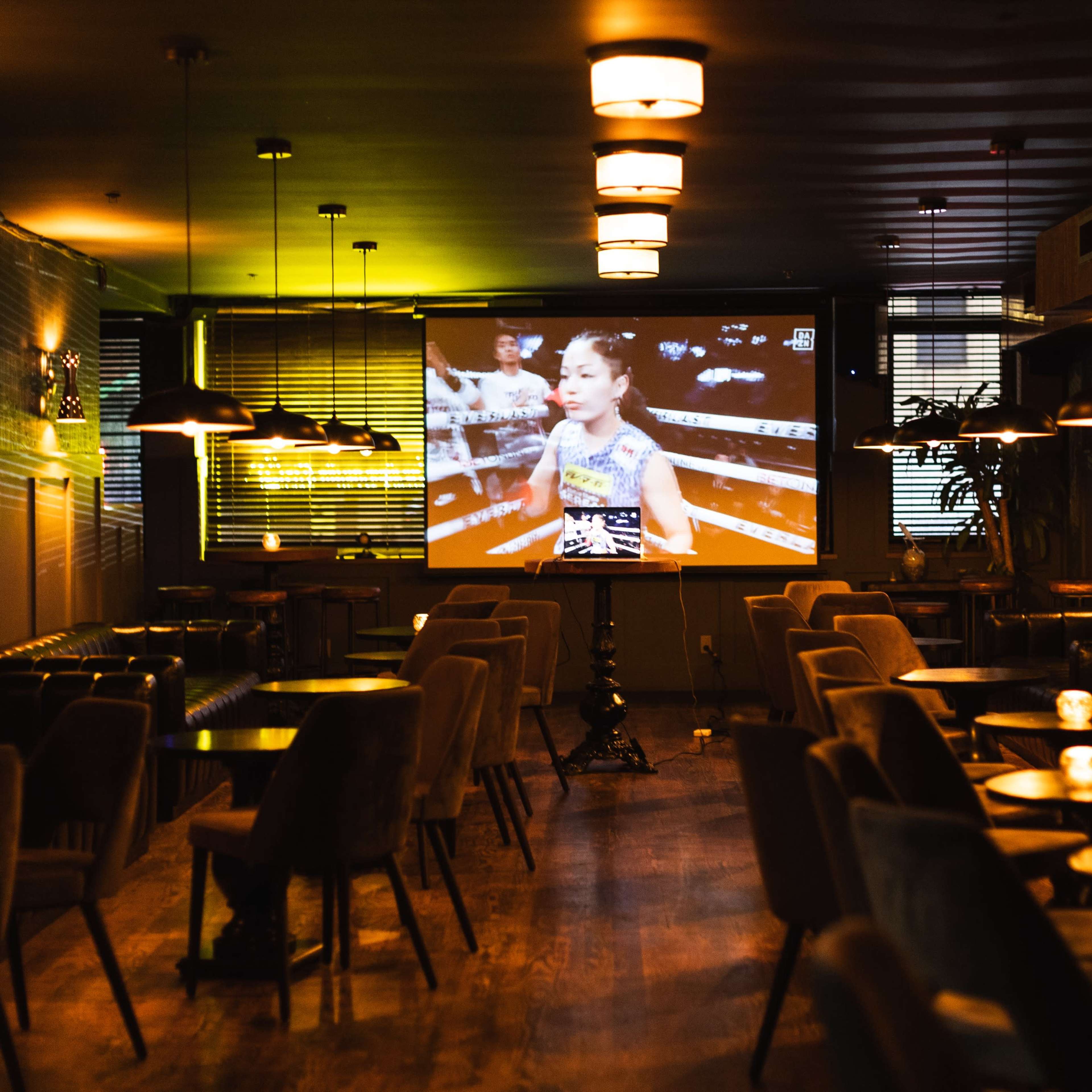 A dimly lit lounge features a large screen displaying a boxing match, with empty tables and chairs arranged around the room.