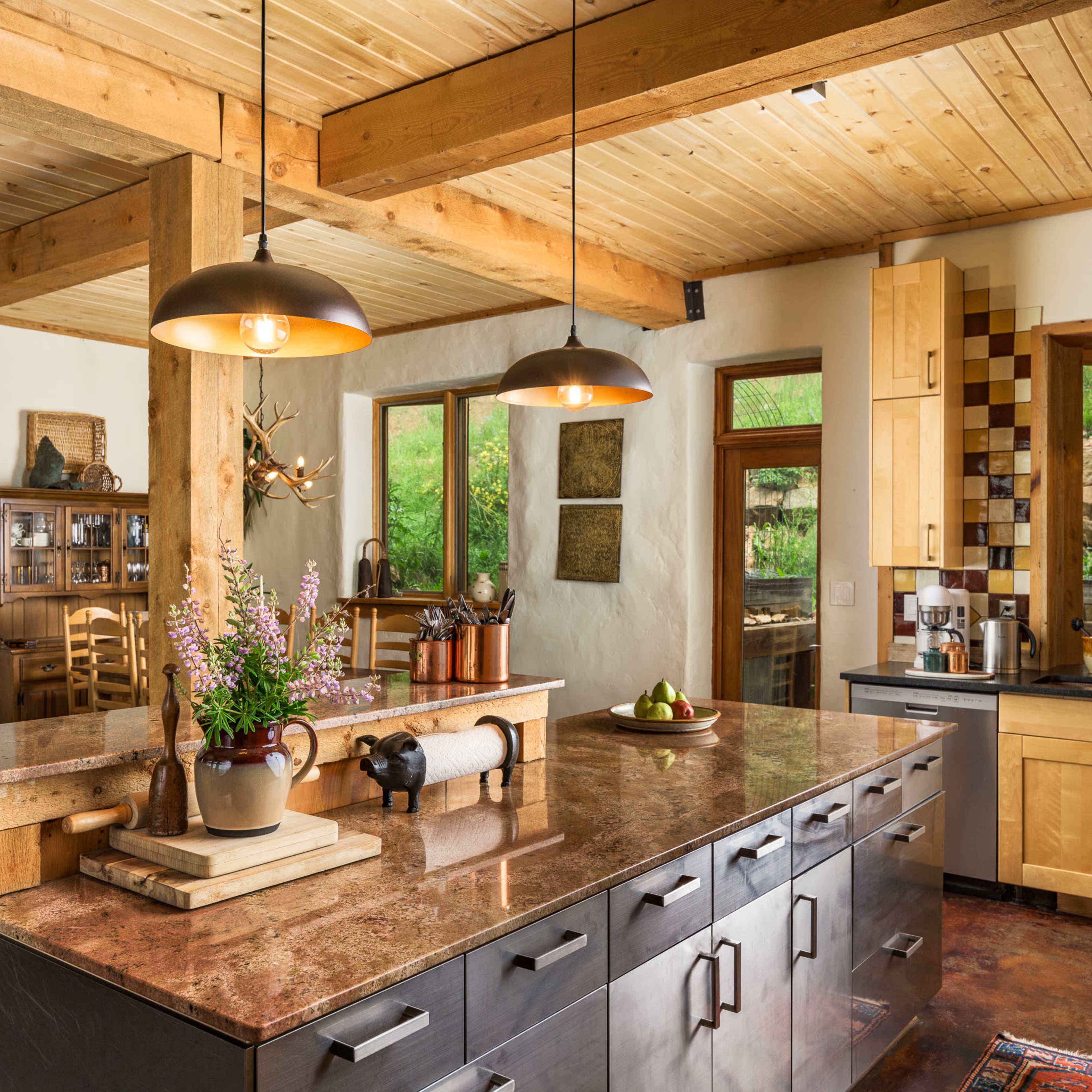 A modern kitchen with wooden beams, a large island with metallic cabinets, and pendant lighting above, complemented by natural elements and large windows.