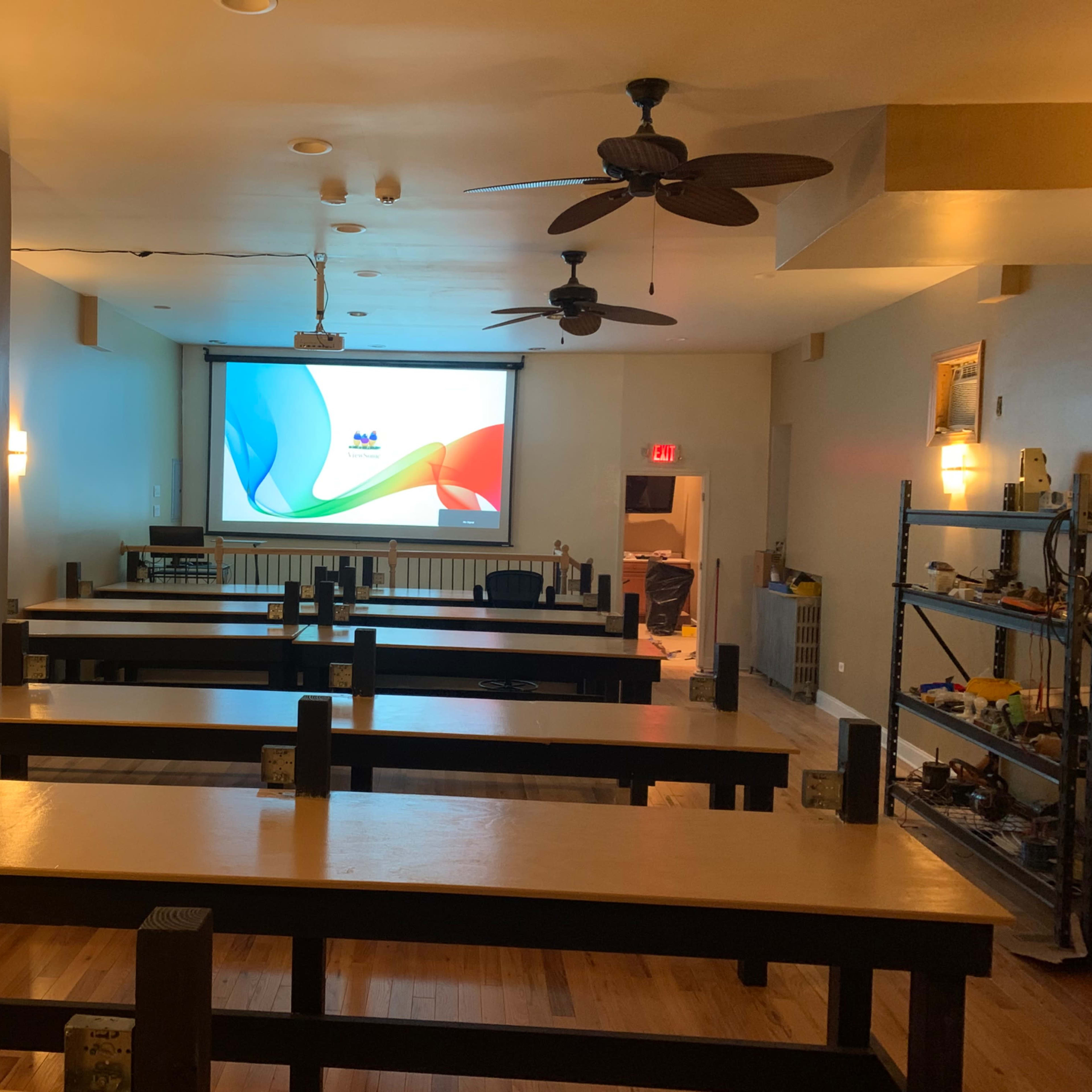An empty room with several long tables, a projector screen on the front wall, and a shelf with various items in the corner.