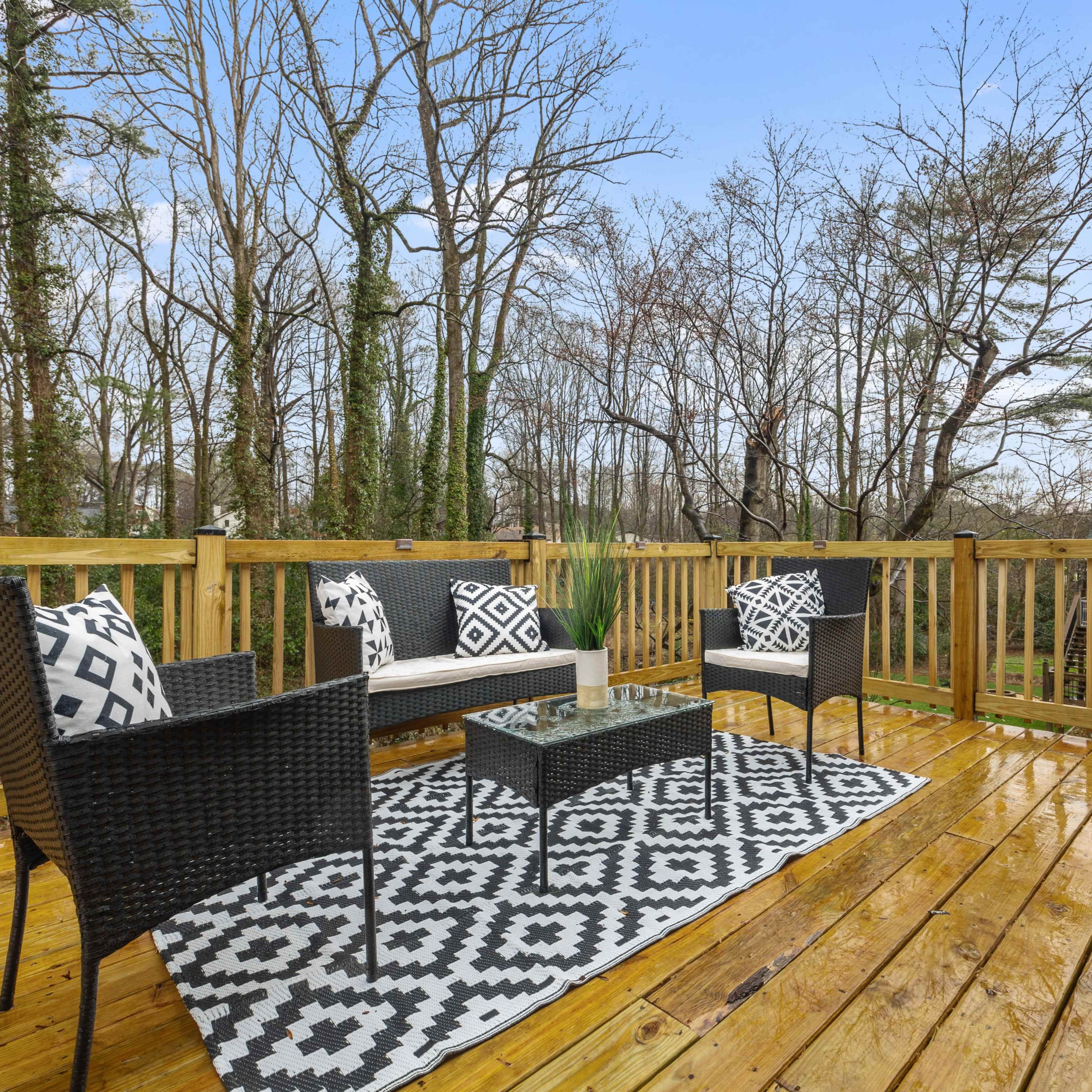 A wooden deck features black and white wicker furniture arranged around a glass coffee table, with a patterned rug and trees visible in the background.