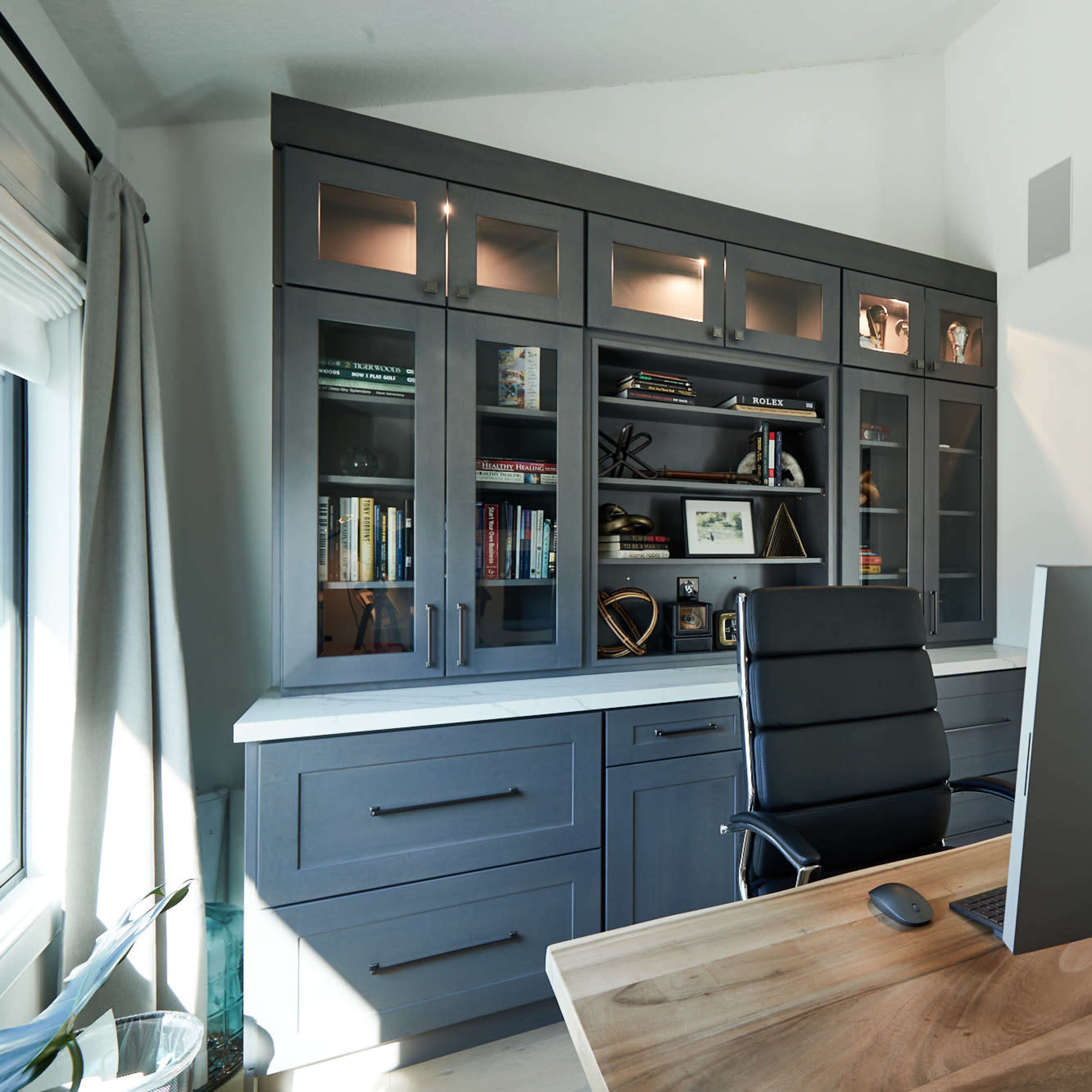 A modern home office with a large desk, an ergonomic chair, and a fitted bookshelf featuring glass cabinets.