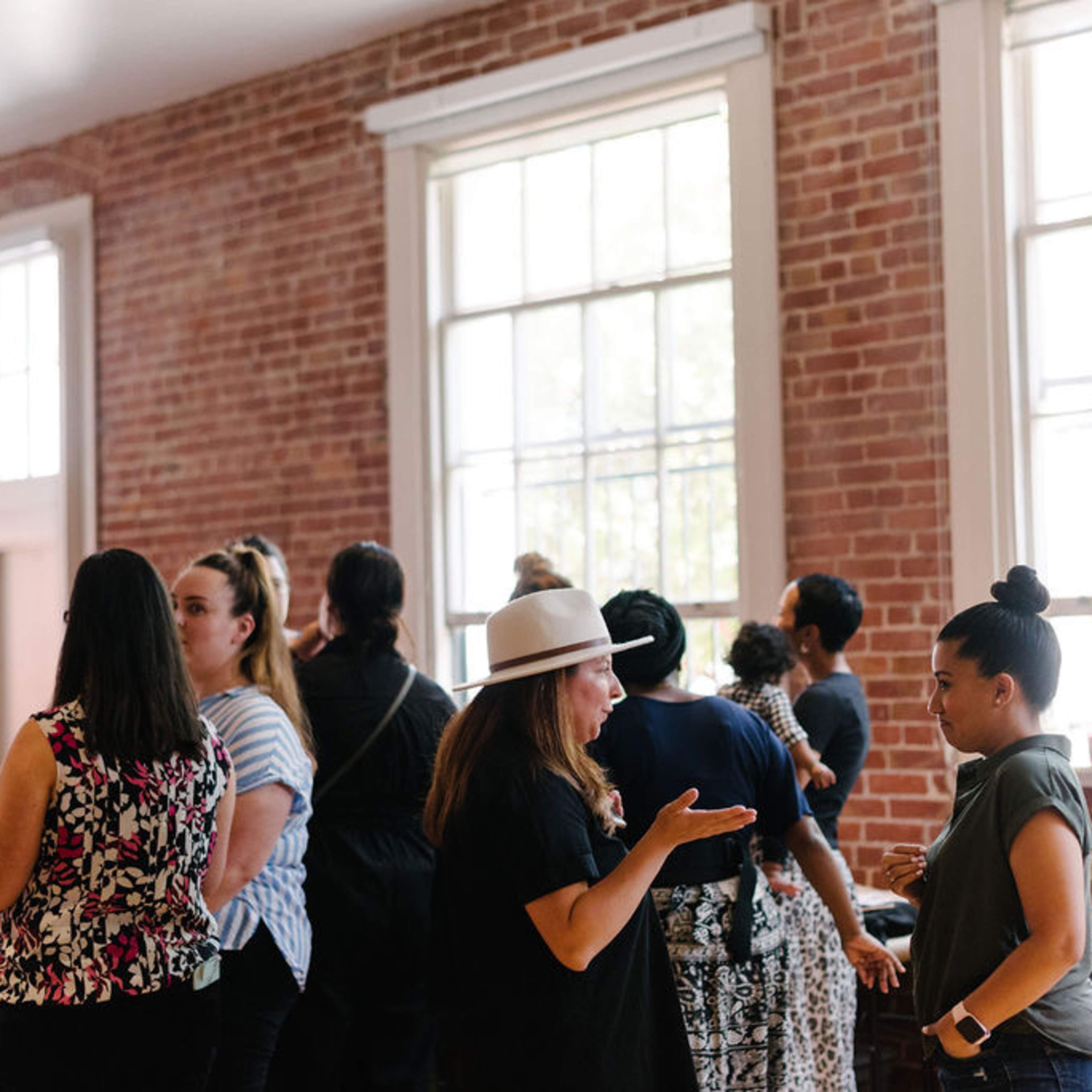 A group of women converse in a spacious room with brick walls and large windows while standing near chairs.