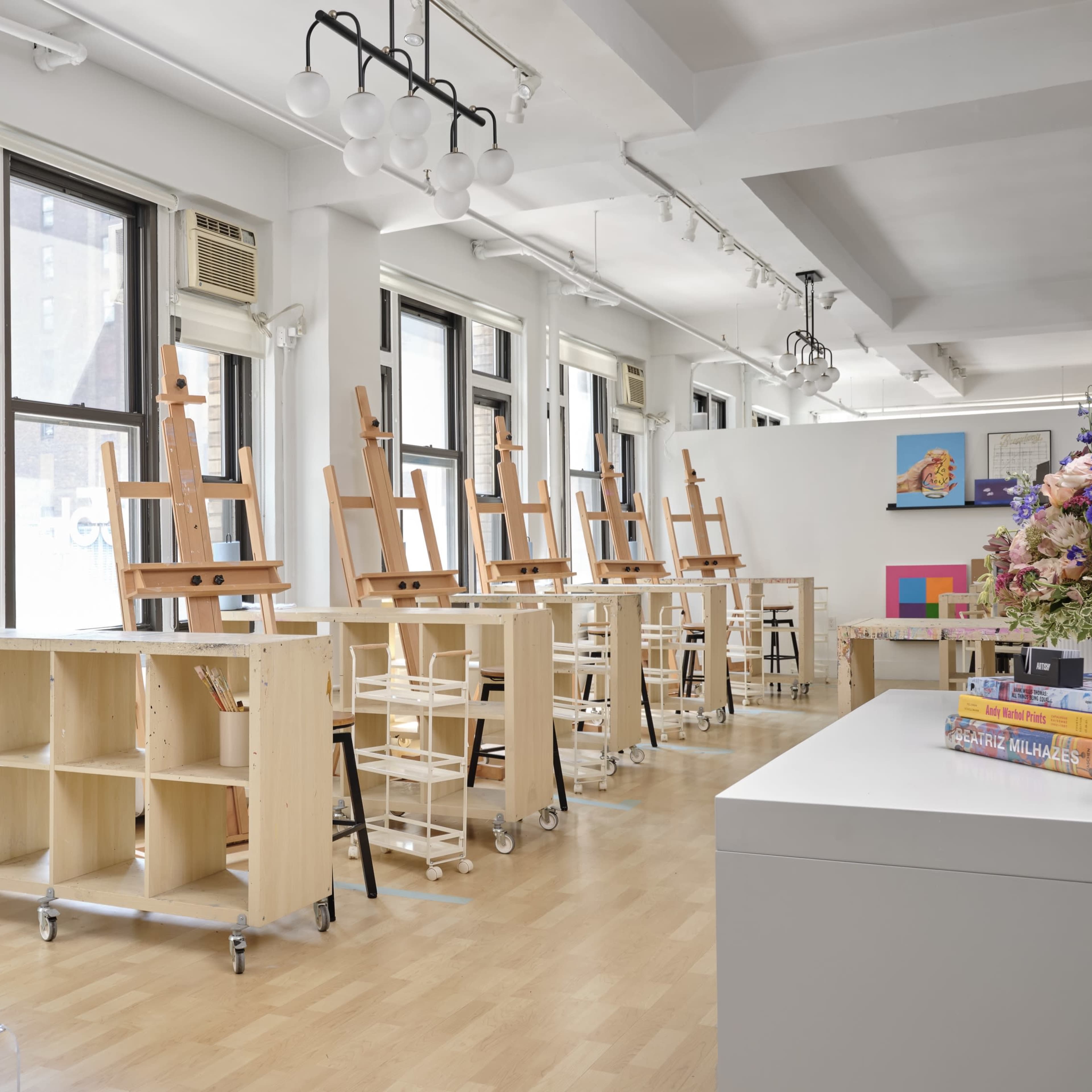 A well-lit art studio featuring several easels and tables arranged in a row beneath large windows.