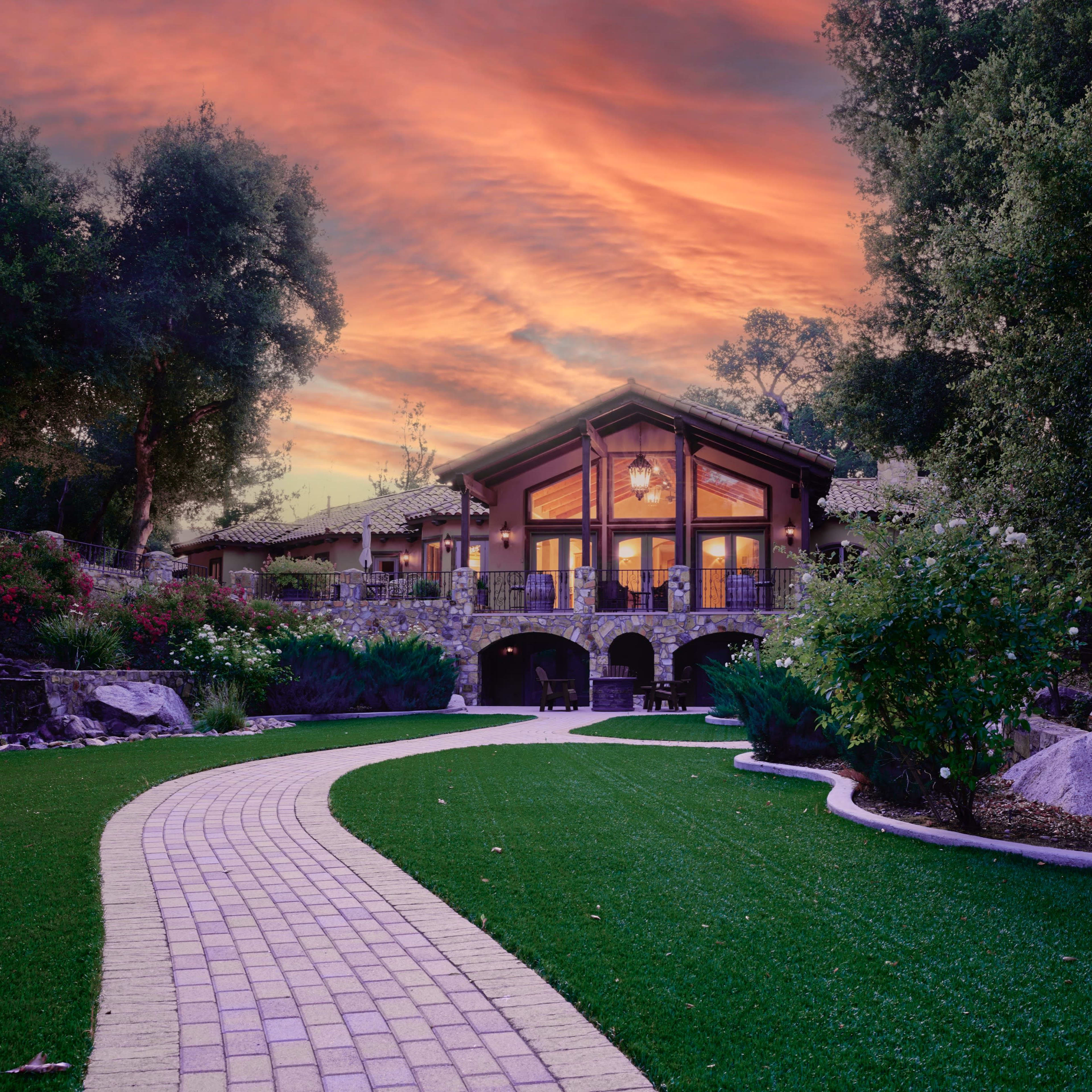 A stone pathway leads to a modern house surrounded by lush greenery and blooming flowers under a colorful sunset.