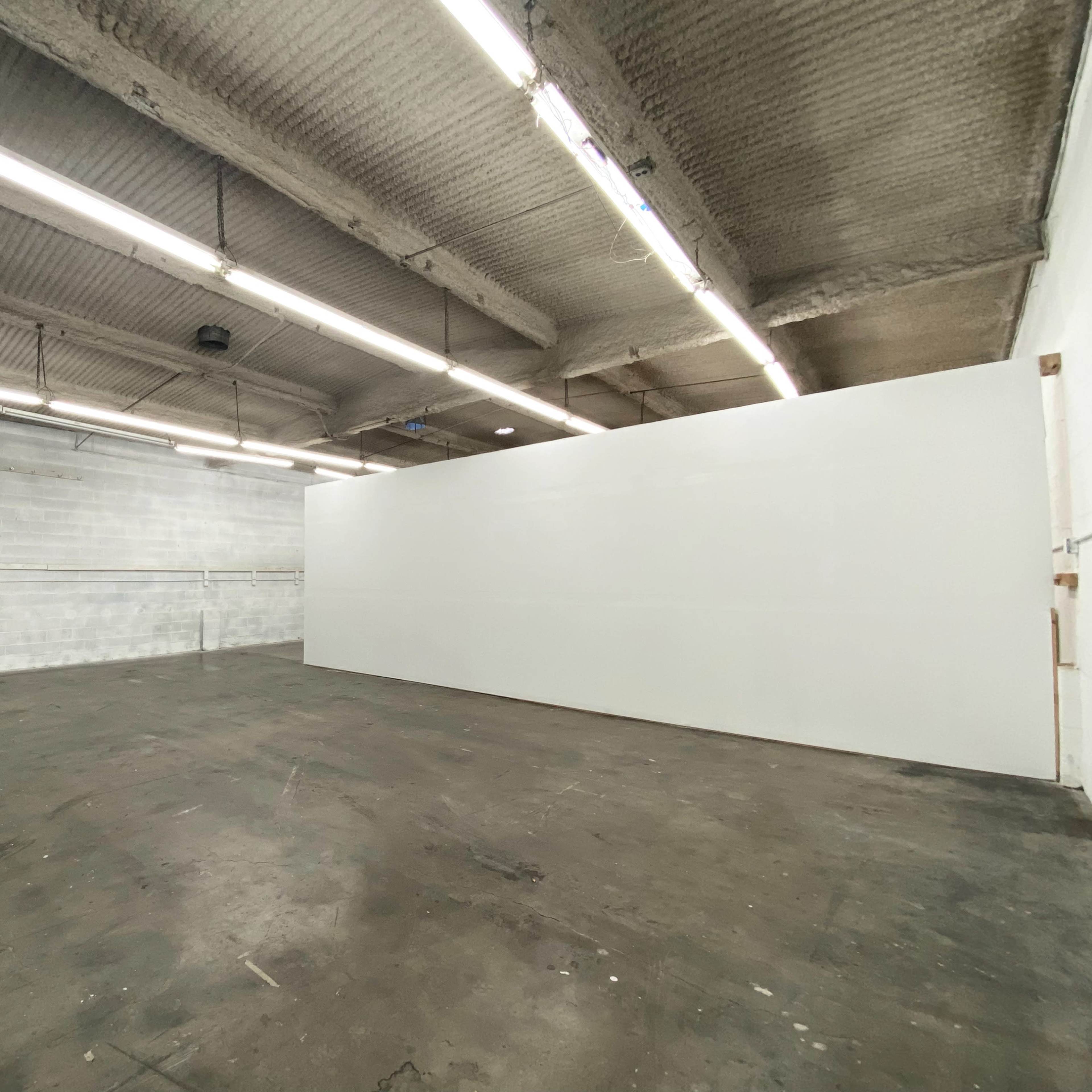 An empty industrial space with white walls, a concrete floor, and overhead fluorescent lighting.