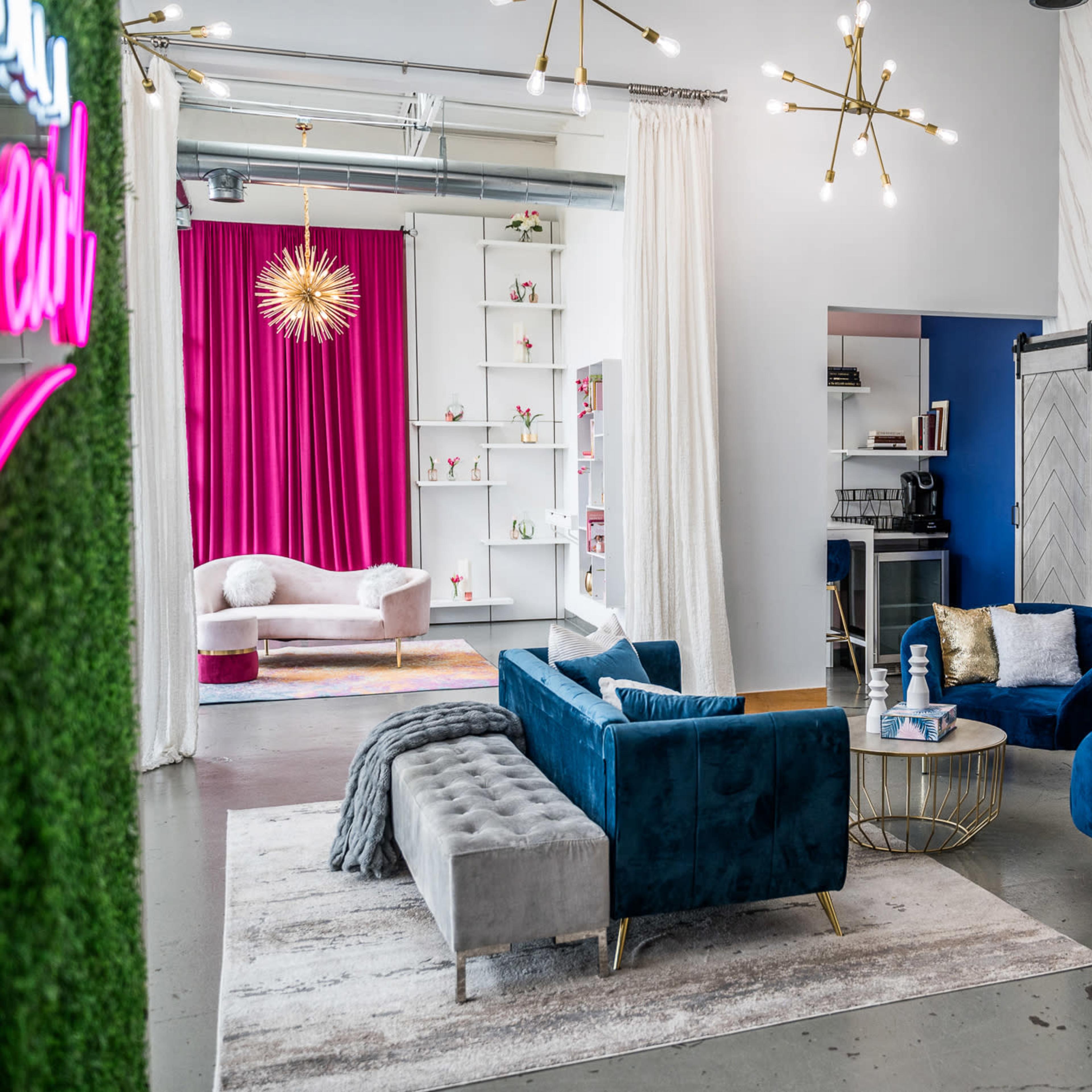 A modern lounge area with blue velvet chairs, a gray tufted sofa, and a decorative table, complemented by a pink backdrop and stylish lighting.
